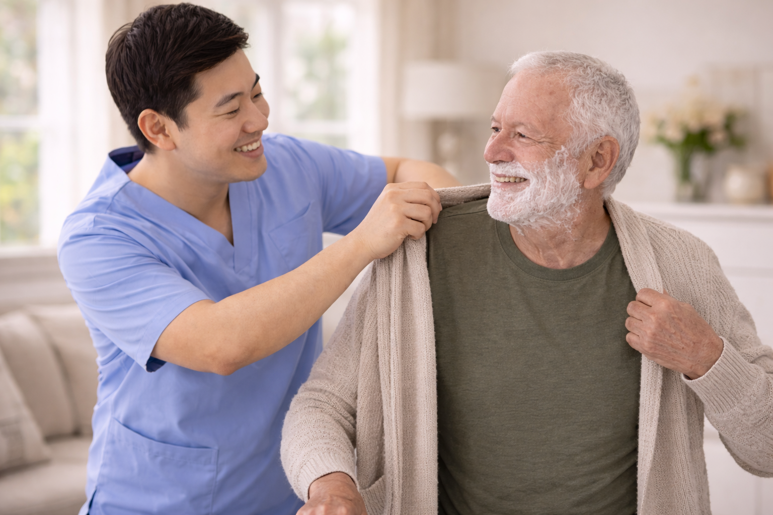 A caregiver helping an elderly man shave by applying shaving cream on his face, both smiling and enjoying the moment in a bright living room.