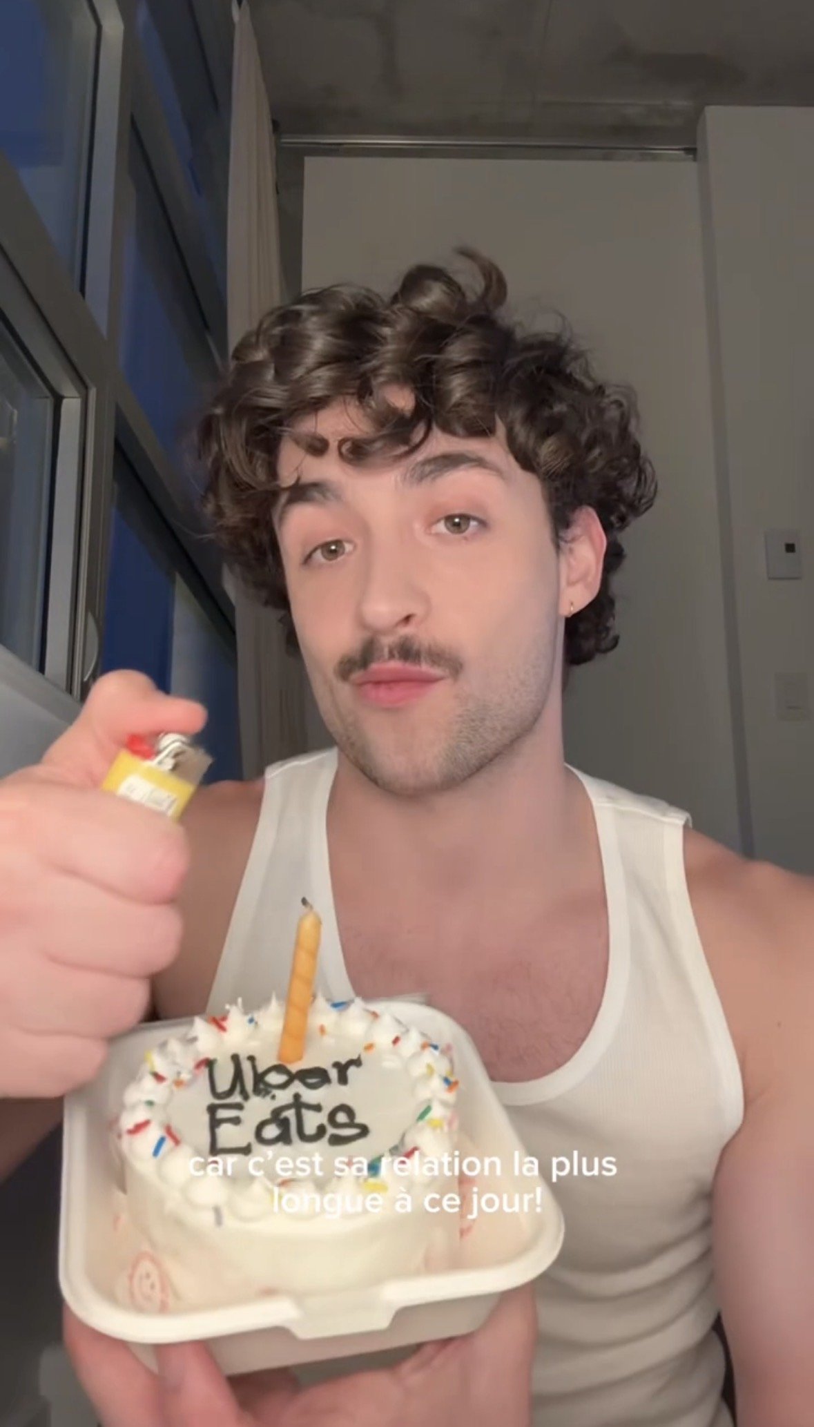 A young man with curly hair and a mustache, wearing a white tank top, holding a birthday cake with a candle on top. The cake has a message written on it and is decorated with colorful sprinkles. The caption in French mentions celebrating a birthday.
