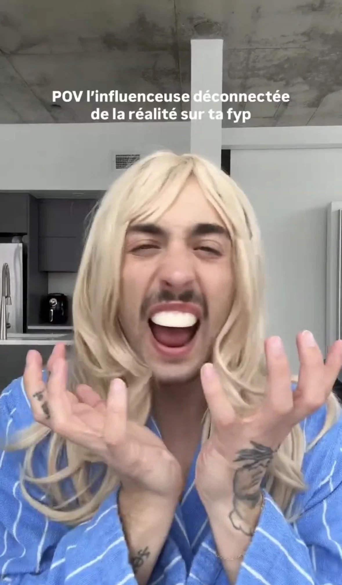 A person with long blonde hair, tattoos, and wearing a blue plaid shirt, smiling with mouth open, showing teeth, and holding their hands near their face, in an indoor setting with gray walls and kitchen appliances in the background.