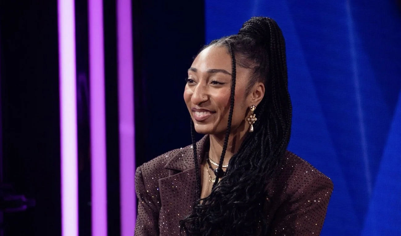 A woman with long braids and jewelry smiling during a televised event or interview.