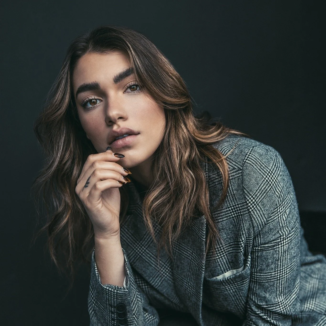 A young woman with wavy brown hair and fair skin posing against a dark background. She is wearing a plaid blazer and has her hand near her chin, with a thoughtful expression.