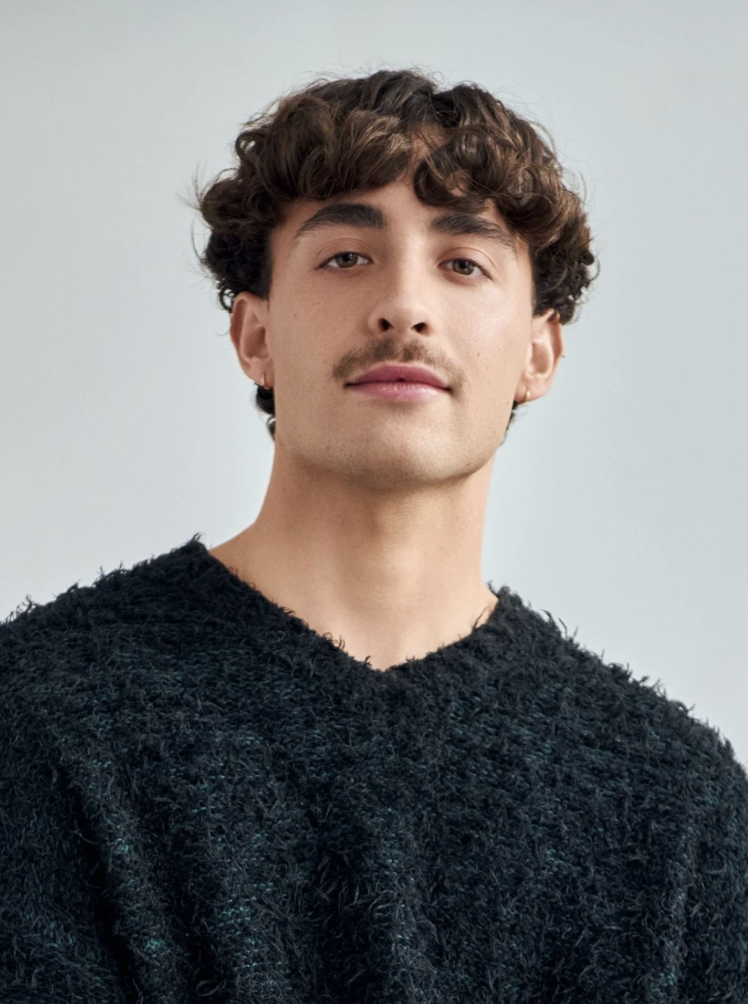 A young man with curly brown hair, light skin, and a thin mustache, wearing a black textured sweater, against a plain light background.