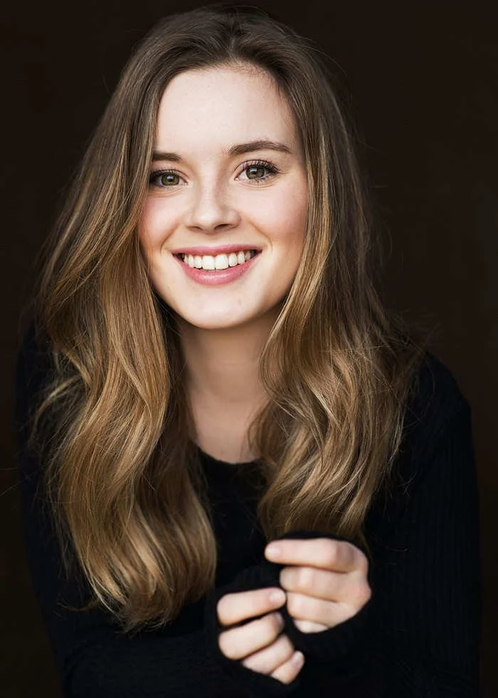 Smiling young woman with long, wavy light brown hair, wearing a black top, against a dark background.
