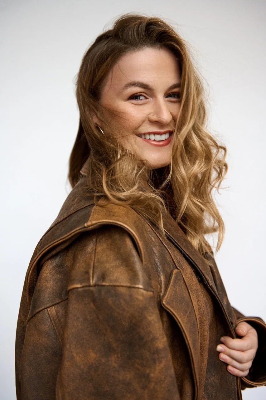 A woman with long, wavy blonde hair smiling and looking at the camera, wearing a brown leather jacket.