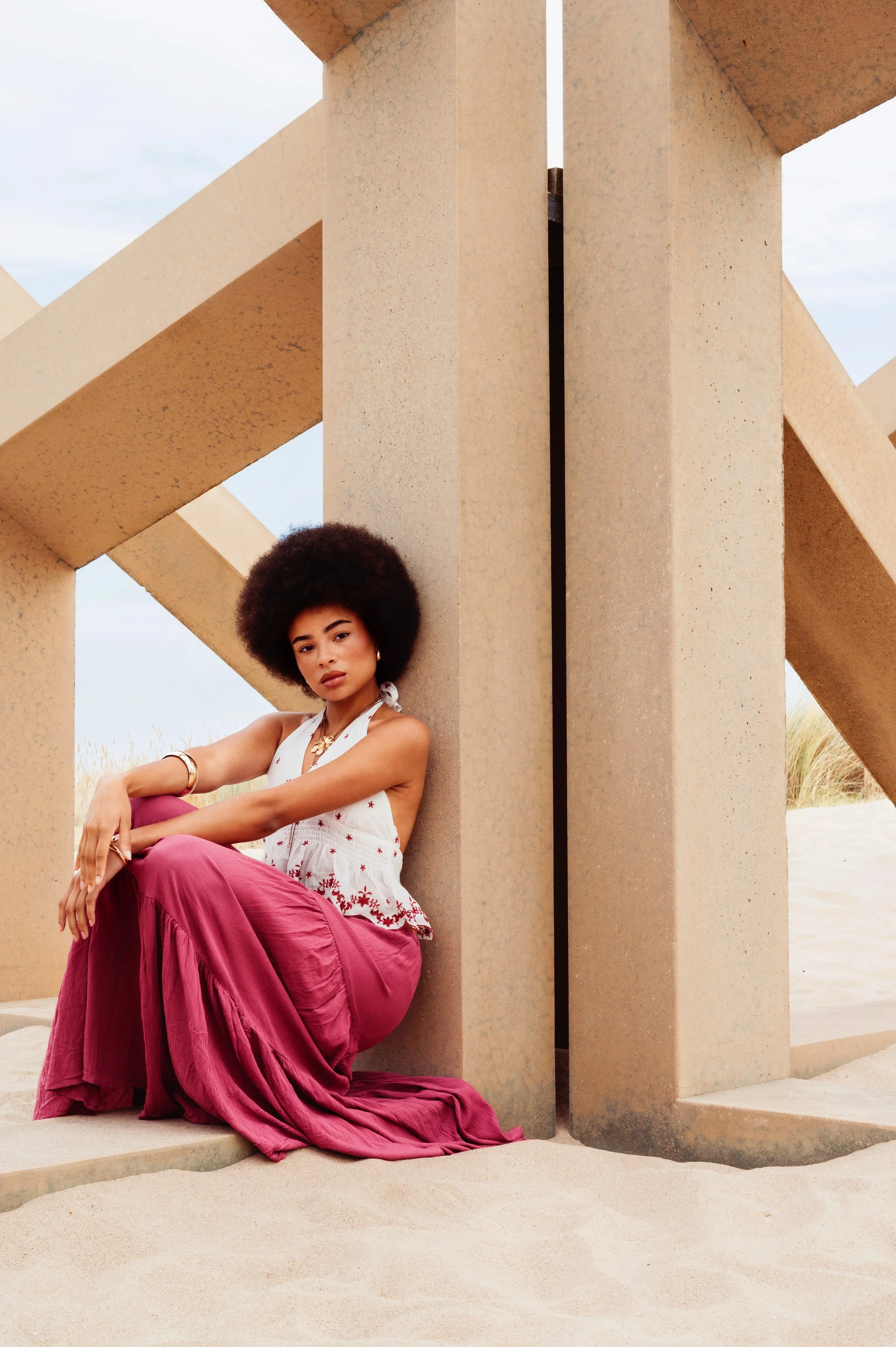 A woman with an afro hairstyle sitting on sand, leaning against a concrete structure at the beach, wearing a white sleeveless top and a long pink skirt.