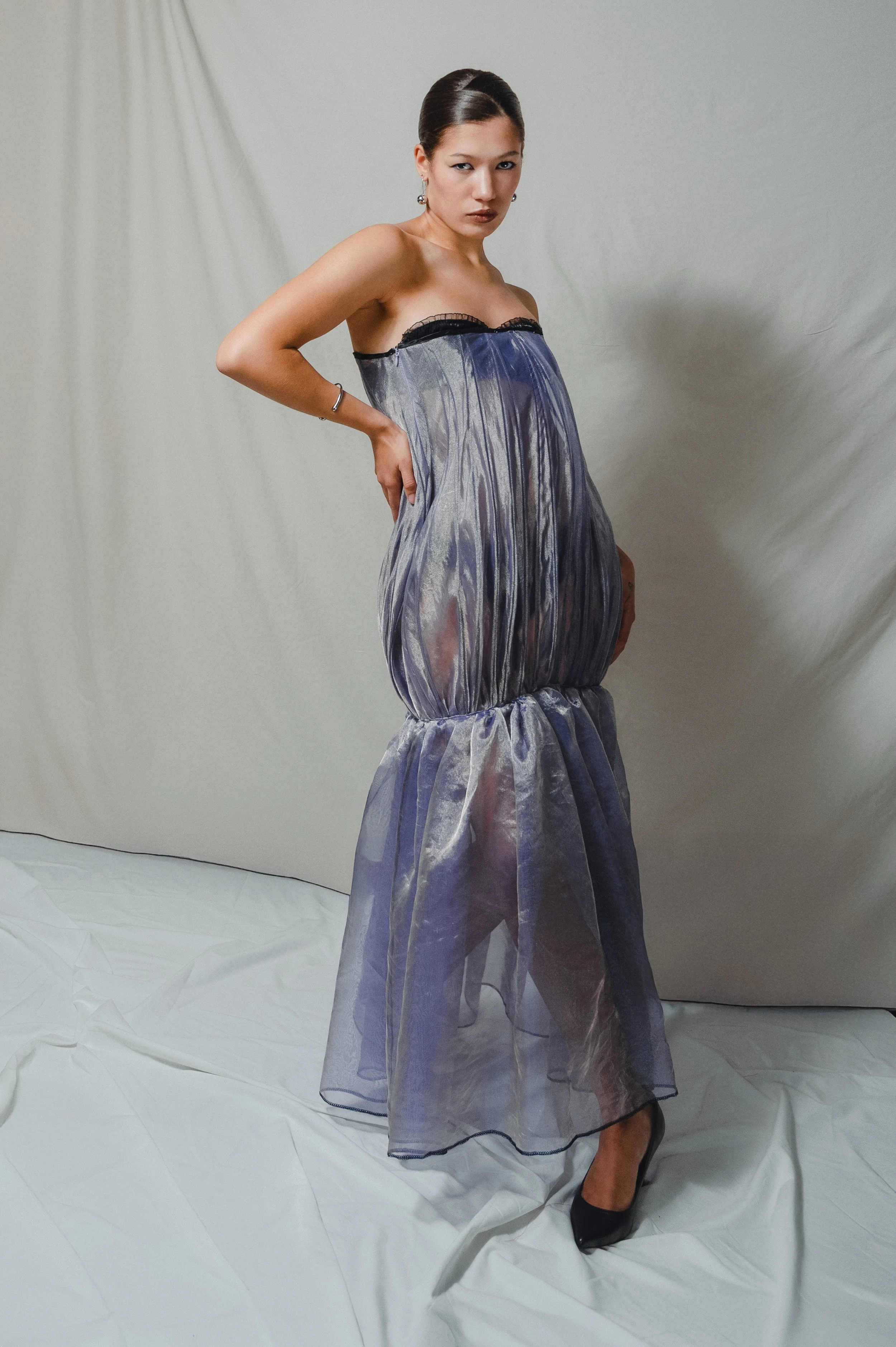 A woman posing in a strapless, sheer, iridescent dress against a plain backdrop.