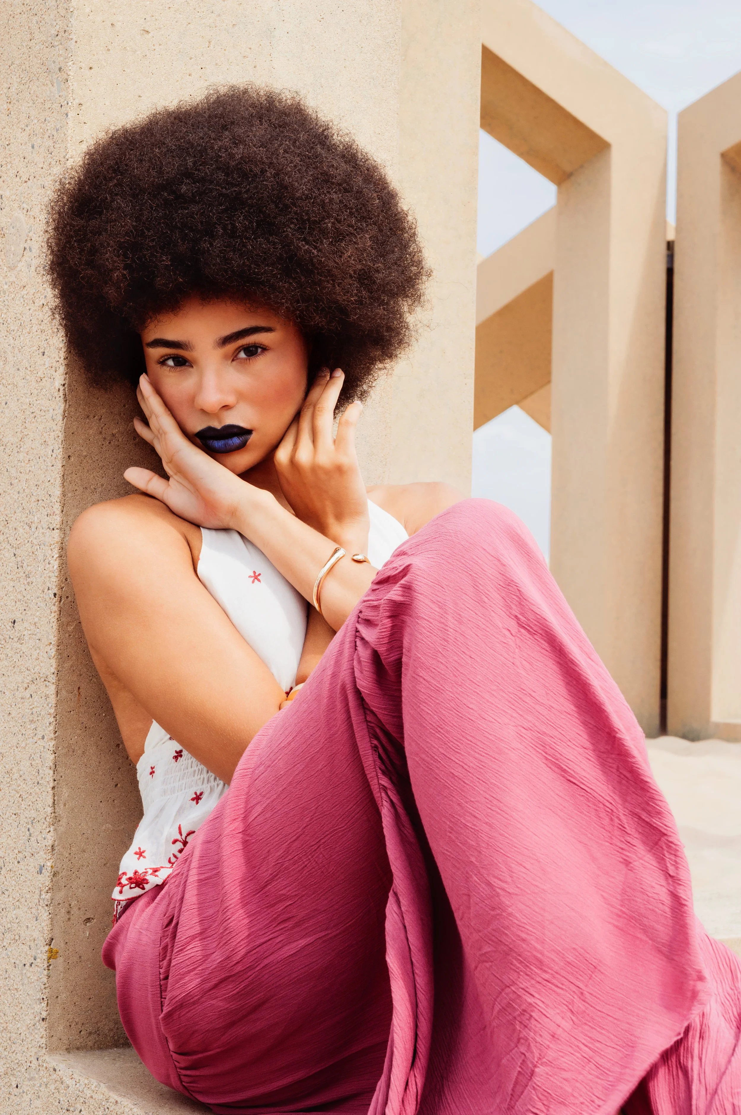 A young woman with an Afro hairstyle and dark lipstick sitting against a concrete wall with architectural structures in the background, wearing a white top and pink pants.