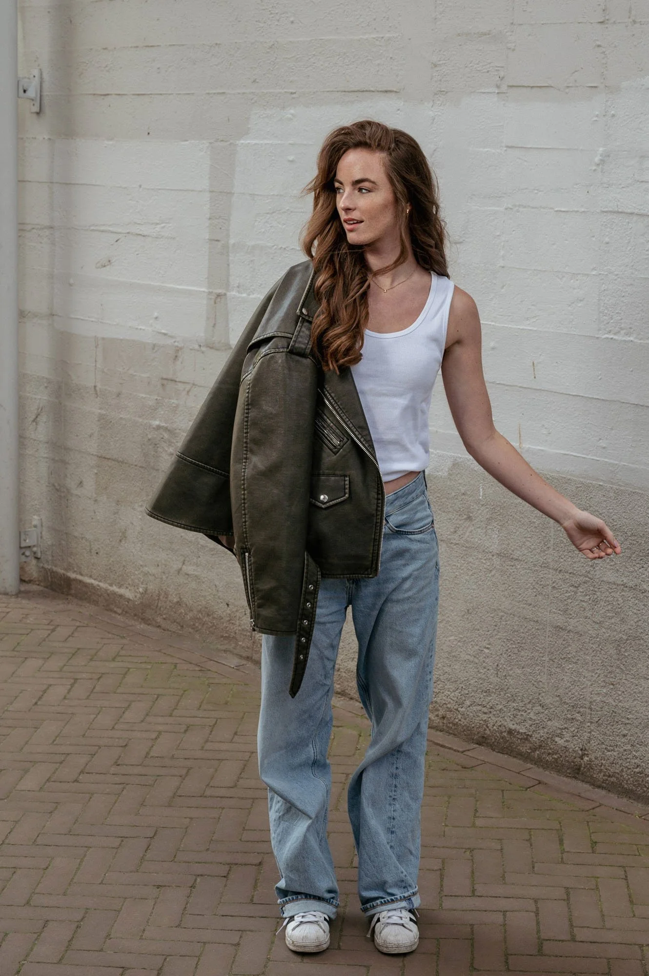 A young woman with long, wavy brown hair, wearing a white tank top, loose jeans, and white sneakers, walking outdoors with a dark green jacket draped over her shoulder, against a brick wall background.