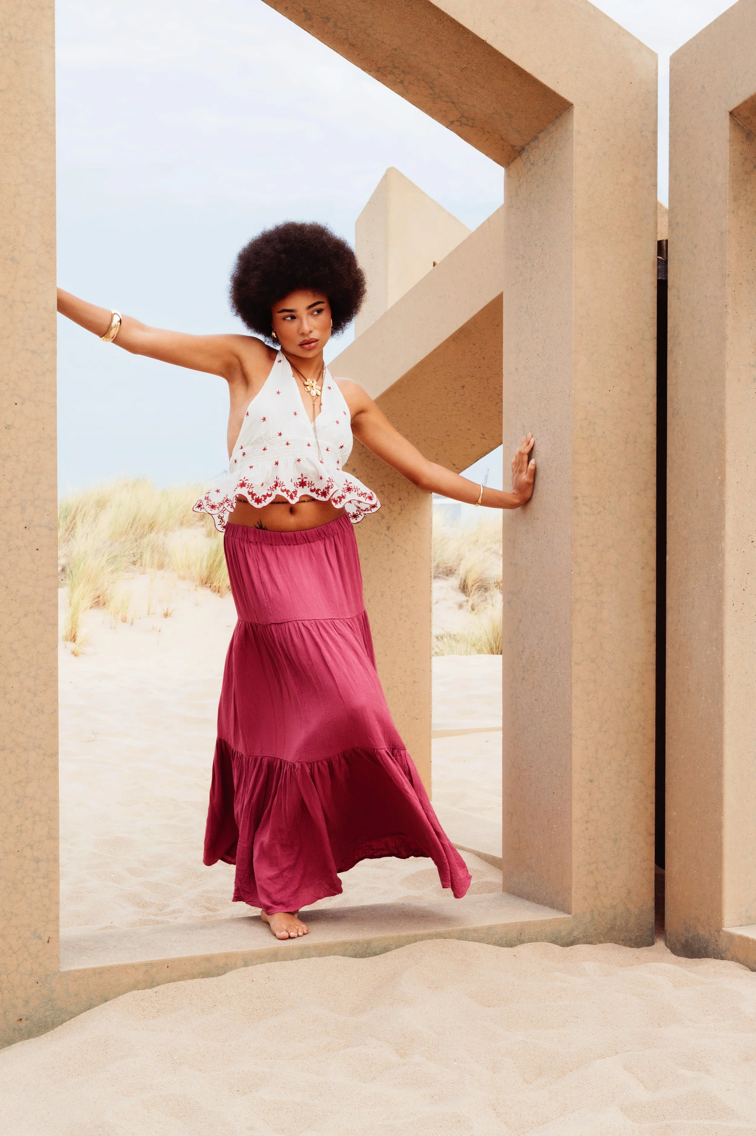 A young woman with an afro hairstyle in a bohemian outfit standing in a modern architectural structure on a sandy beach, with dunes and grasses in the background.