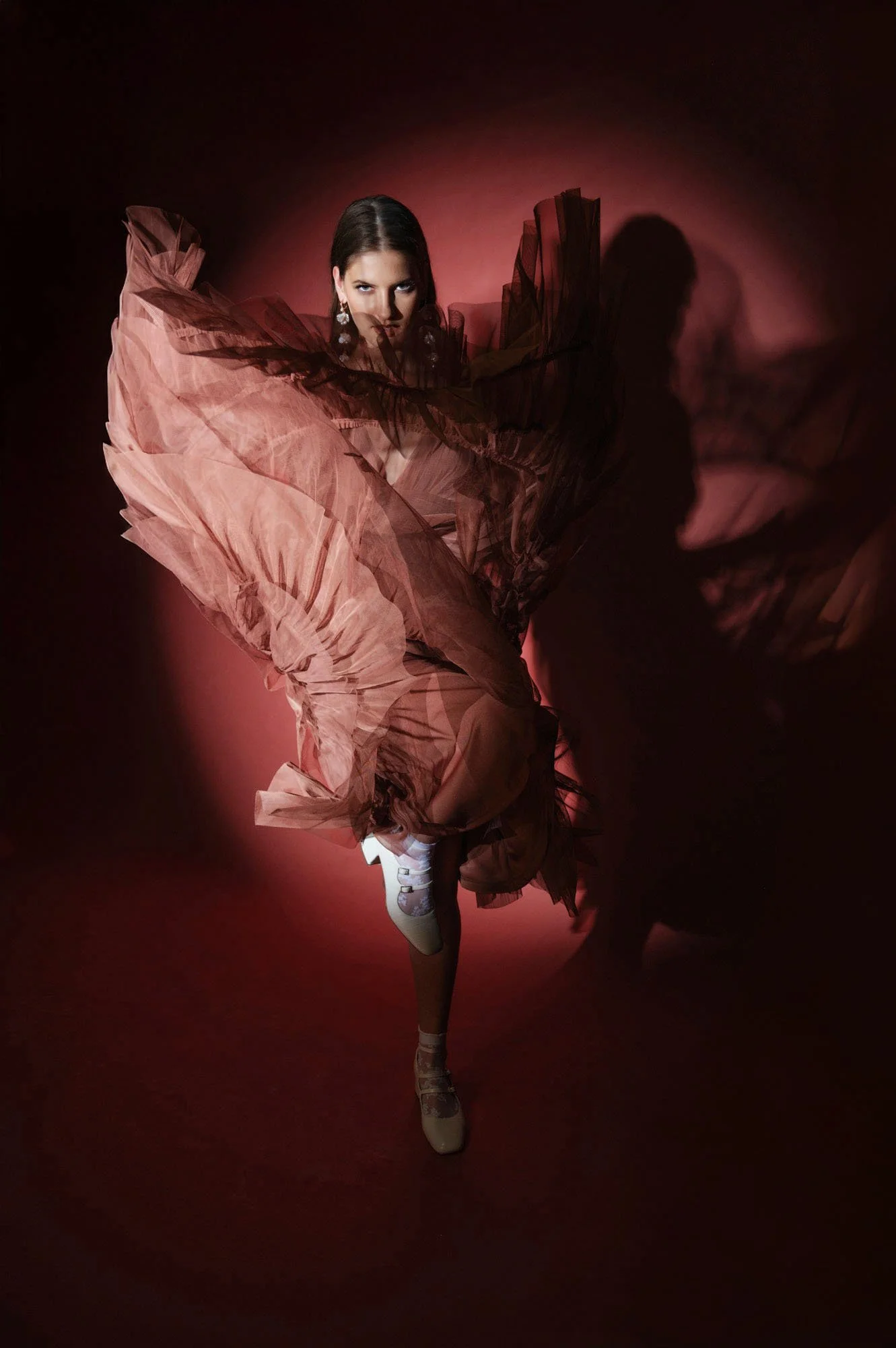 A woman in a flowing, layered, pink and brown dress with dramatic sleeves, posing against a pink background with a strong shadow cast on the wall.