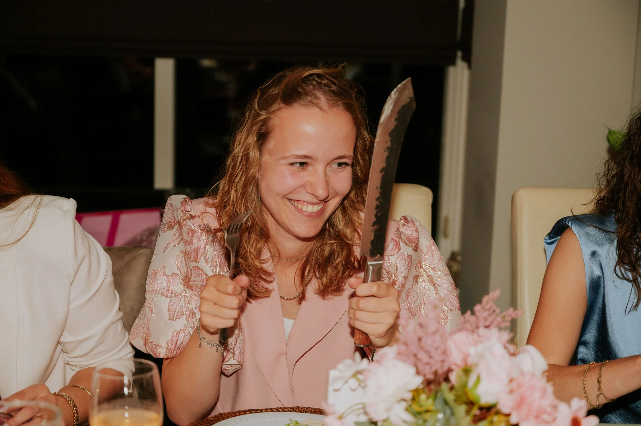 Woman with curly red hair holding a large kitchen knife in one hand and a fork in the other, smiling at a dinner table with floral centerpiece during a gathering.