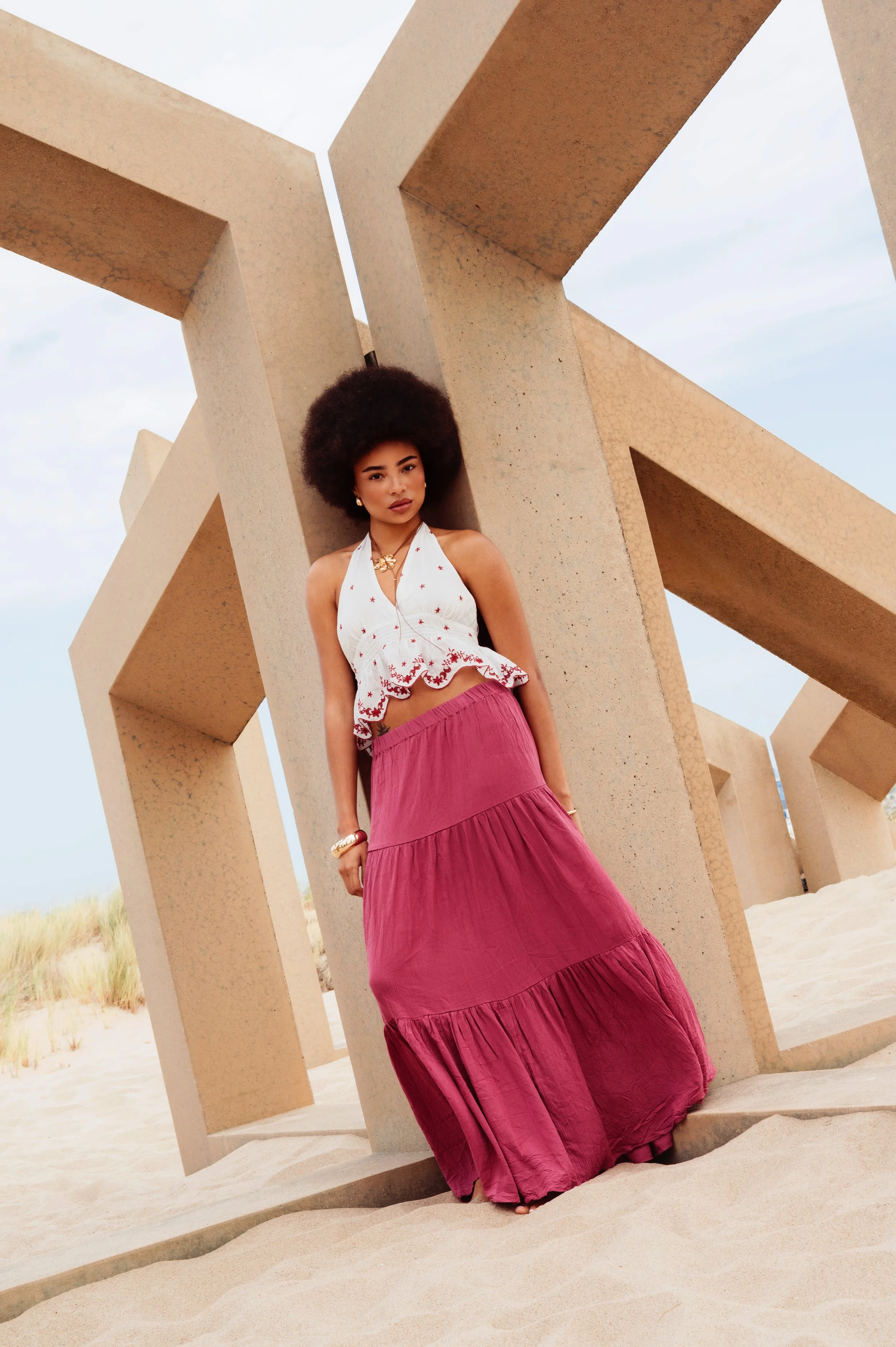 A young woman standing on a sandy beach, leaning against a large concrete sculpture with geometric shapes, wearing a white halter top and a long pink skirt, with curly afro hair and jewelry.