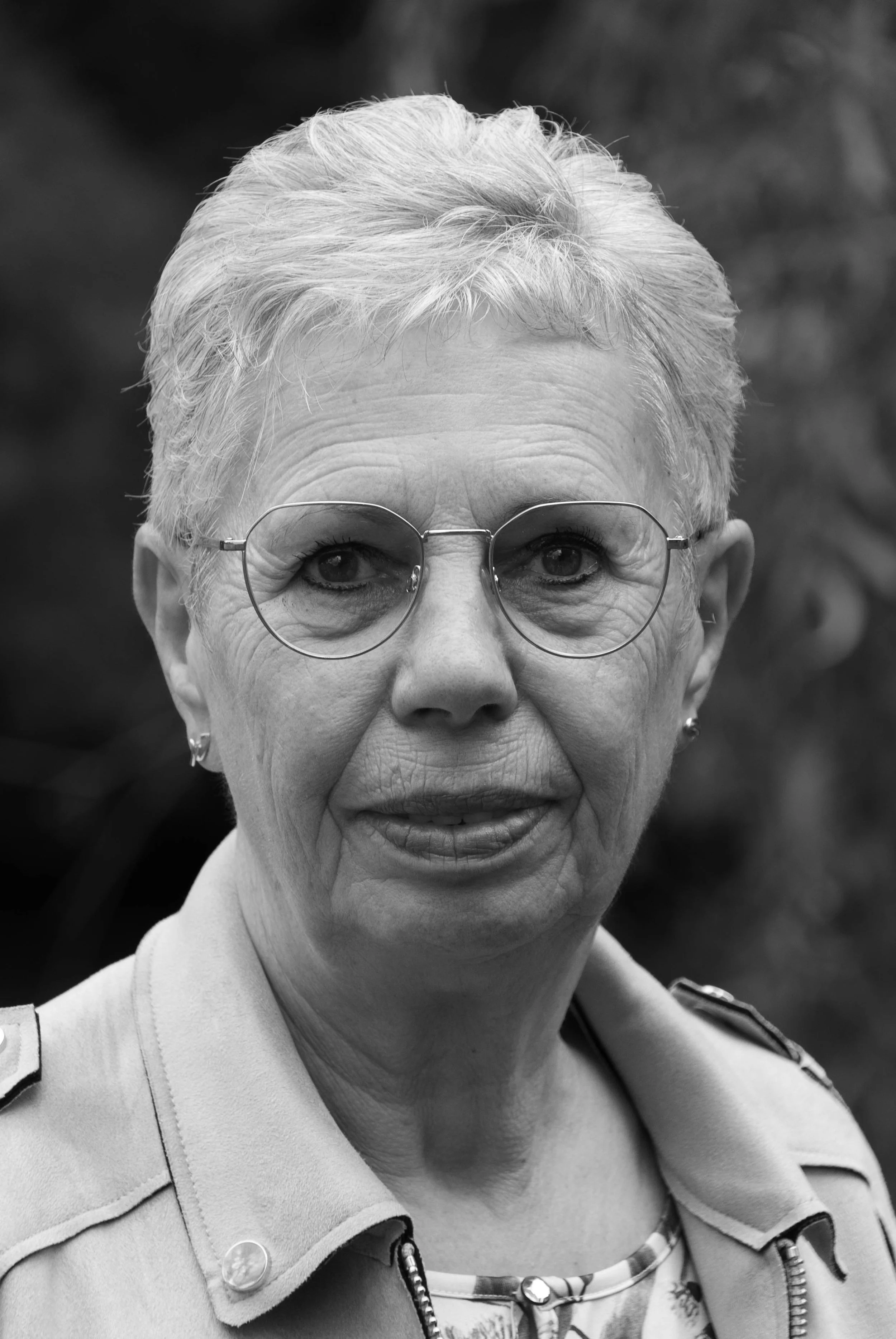 A black and white portrait of an older woman with short, light-colored hair, wearing glasses, earrings, and a jacket with a zipper and buttons.