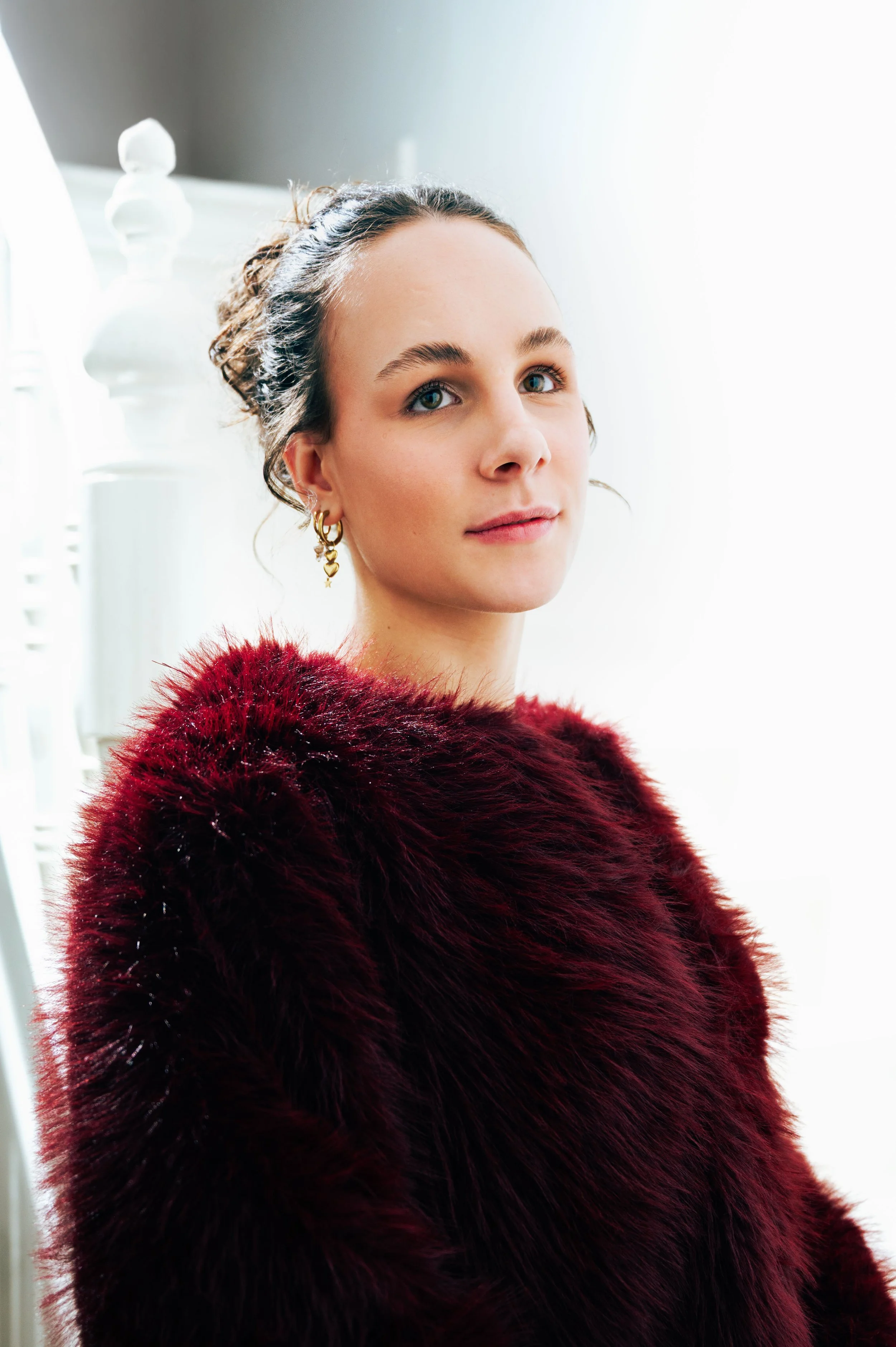 Portrait of a young woman with light skin and blue eyes wearing a burgundy fur coat and gold earrings, standing indoors near a white door with sunlight coming through.