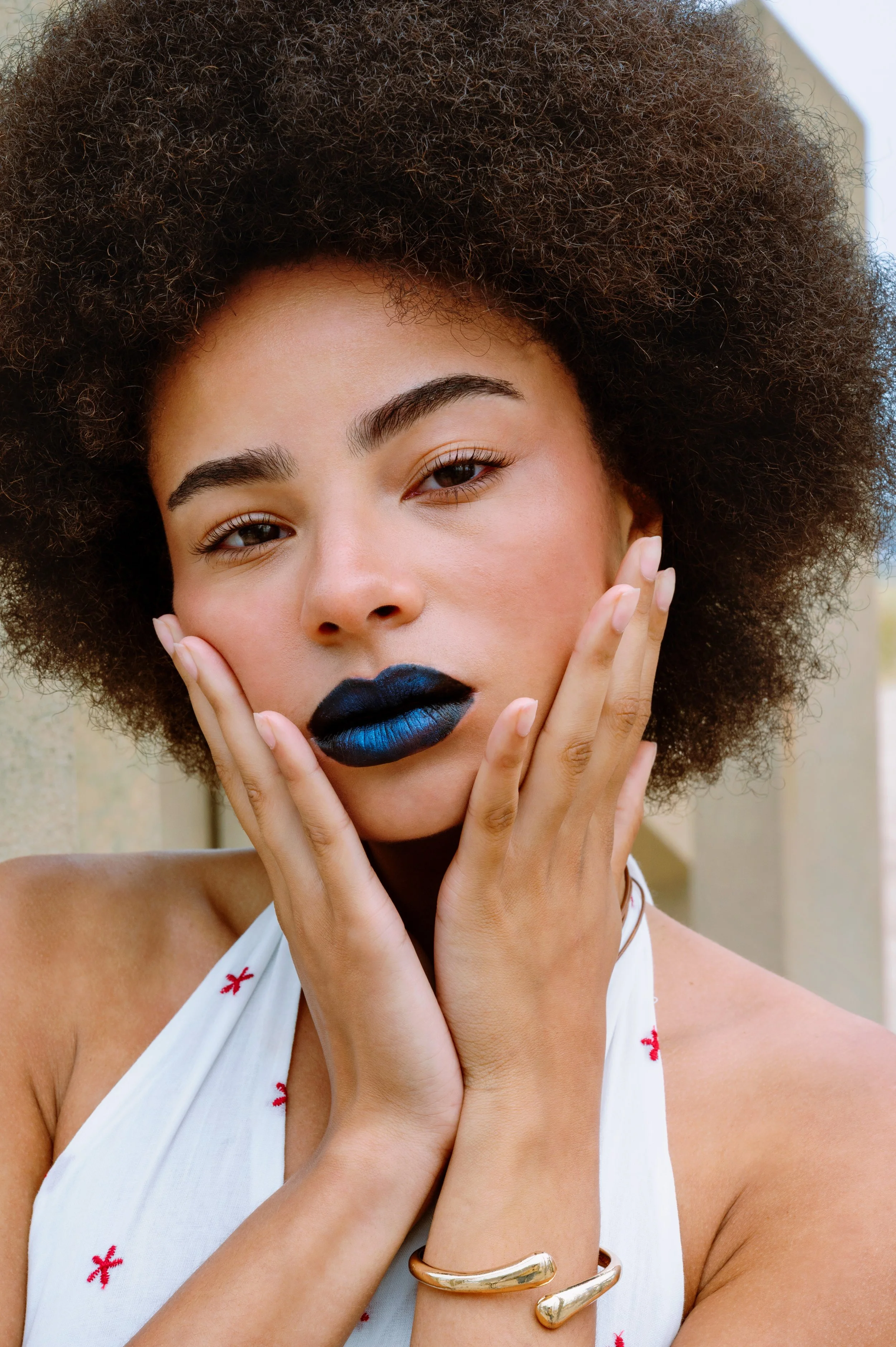 A woman with curly hair and natural makeup, wearing a white sleeveless top with small red embroidery, has her hands on her face. She has striking black and blue ombre lipstick and is outdoors, with a blurred background.