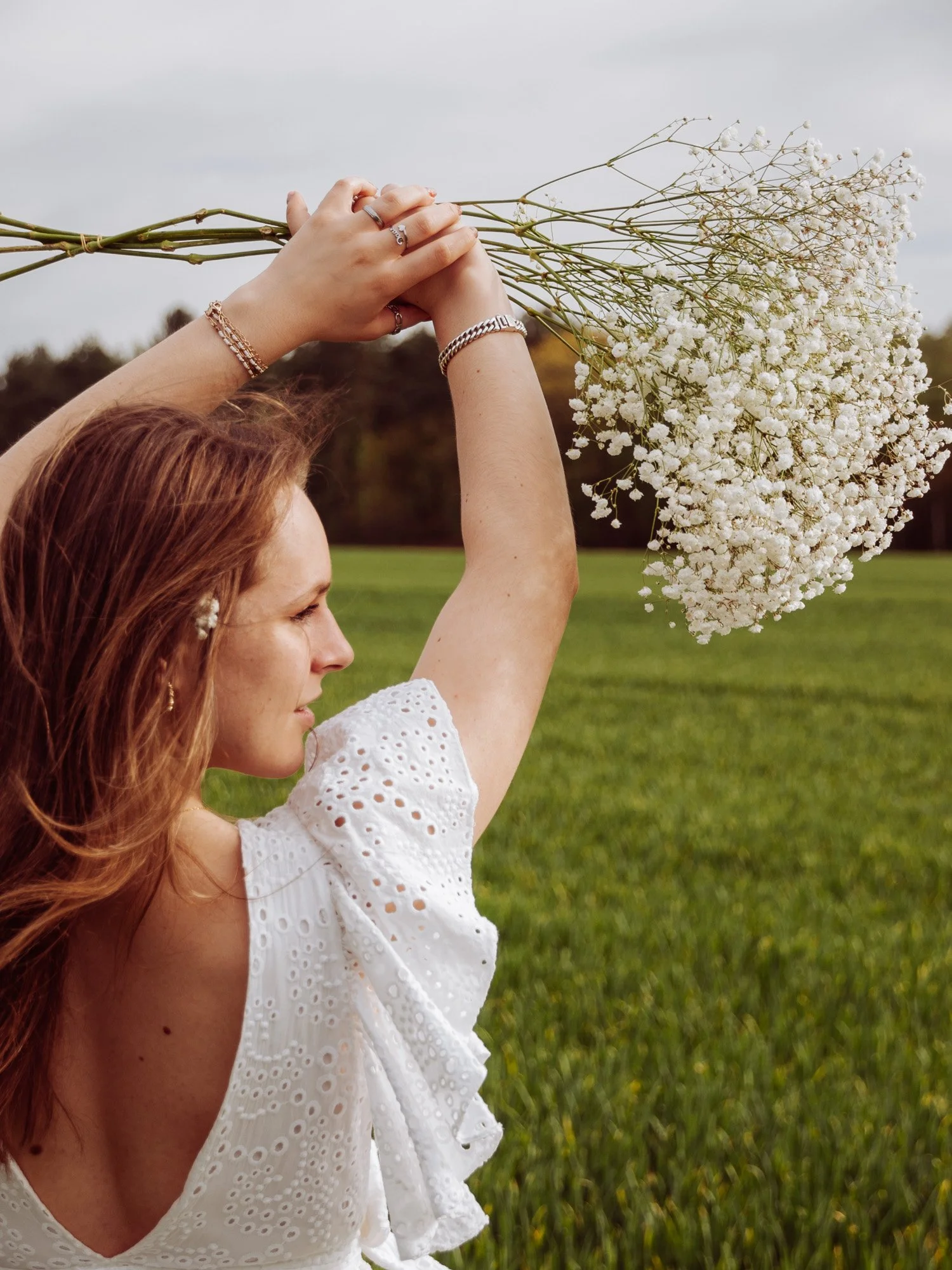 A woman with red hair in a white eyelet dress holds a bunch of white baby's breath flowers above her head in an outdoor grassy field.