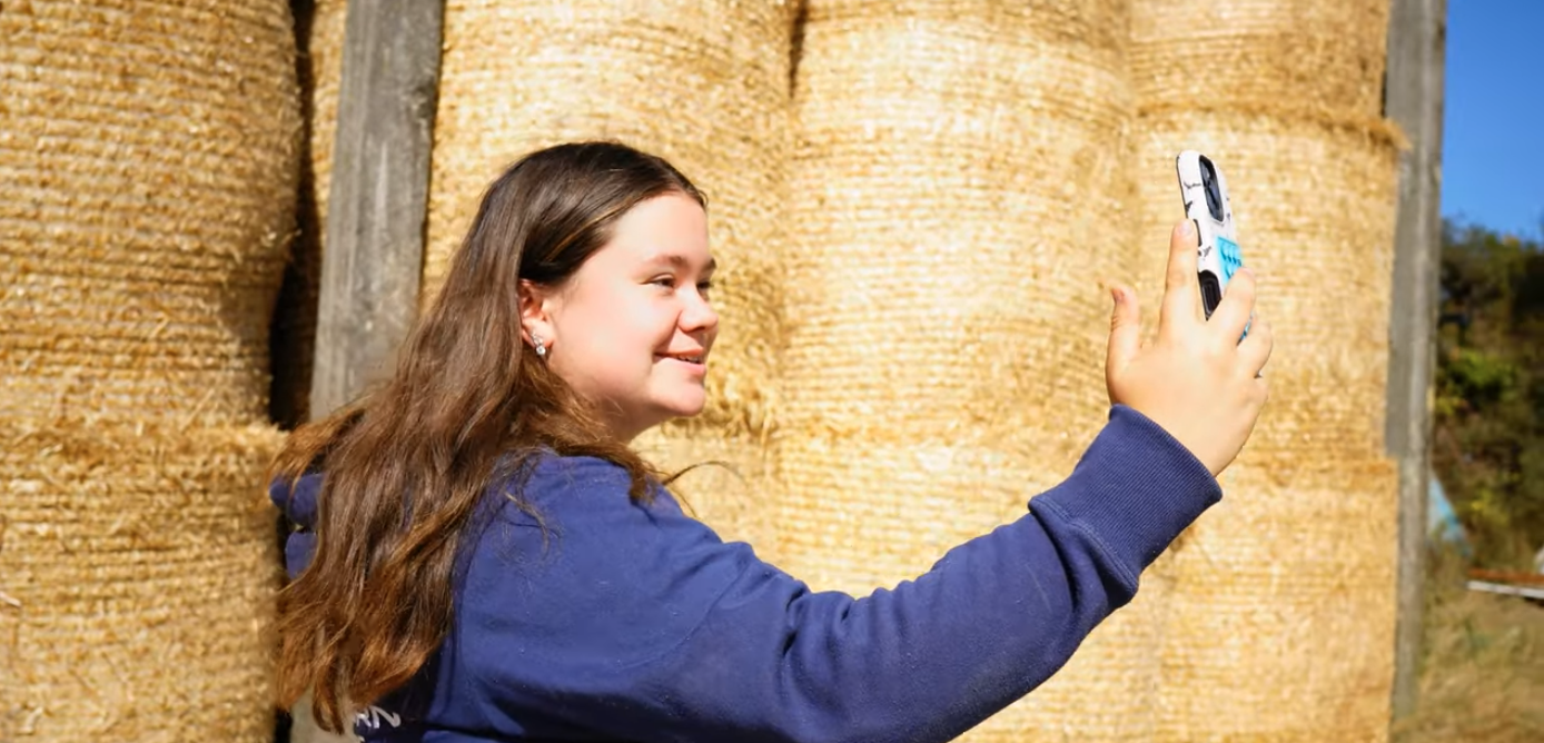Young girl taking a selfie in front of a large hay bale with a handheld device.