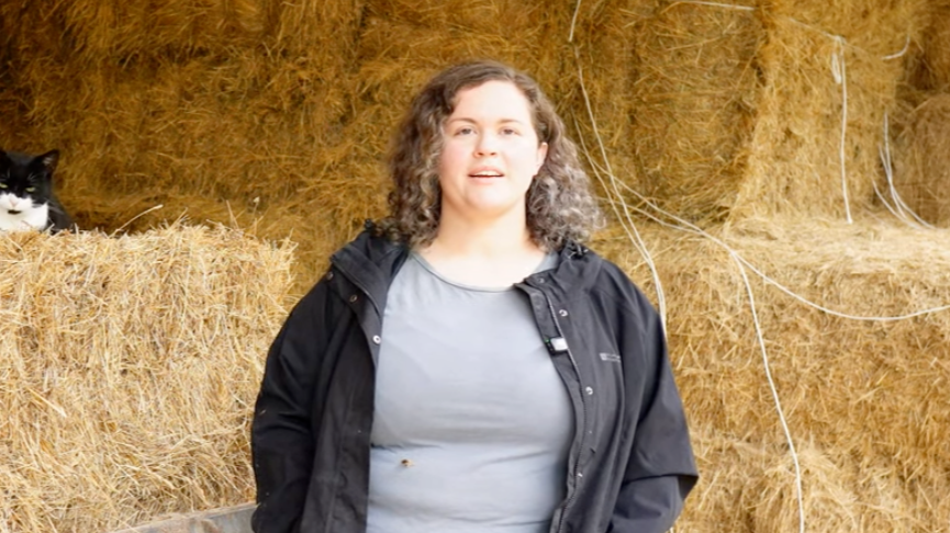 A woman standing in front of a backdrop of stacked hay bales, with a black and white cat partially visible on the left side.