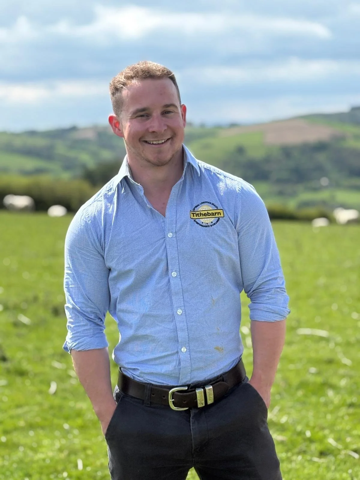 A young man with short, light brown hair, wearing a light blue button-up shirt with a 'Tithebarn' logo on the chest, standing outdoors in a green field with a rural landscape and hills in the background on a cloudy day, smiling at the camera.