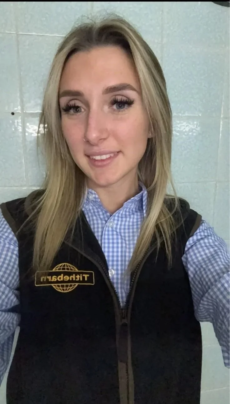 A young woman with blonde hair, wearing a checkered blue shirt and a black vest with a 'Tithebarn' logo, taking a selfie in front of a tiled wall.