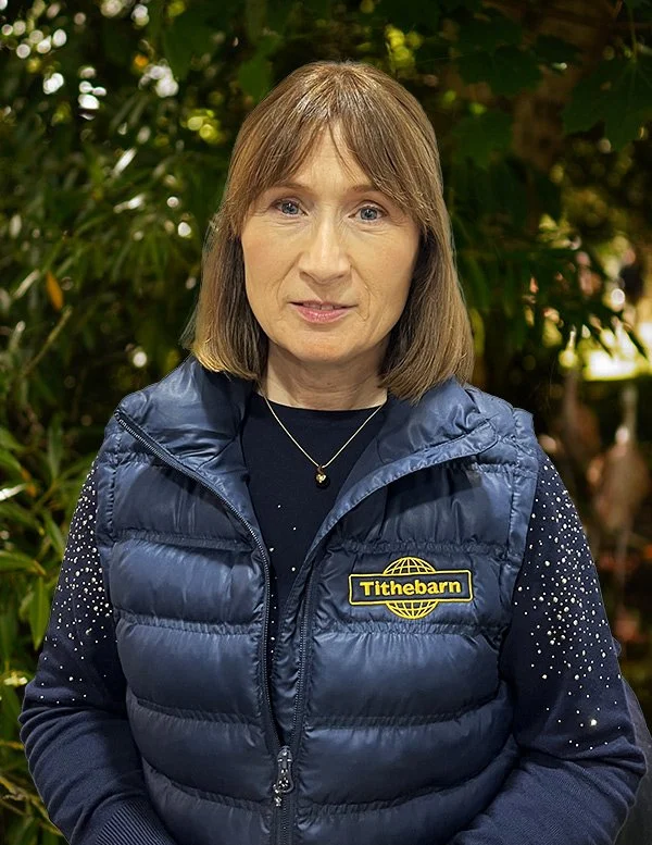 A woman with shoulder-length light brown hair and light skin standing outdoors in front of green foliage, wearing a navy blue puffer vest with a yellow and black 'Tithebarn' logo, a dark long-sleeve shirt with small white dots, and a gold necklace.