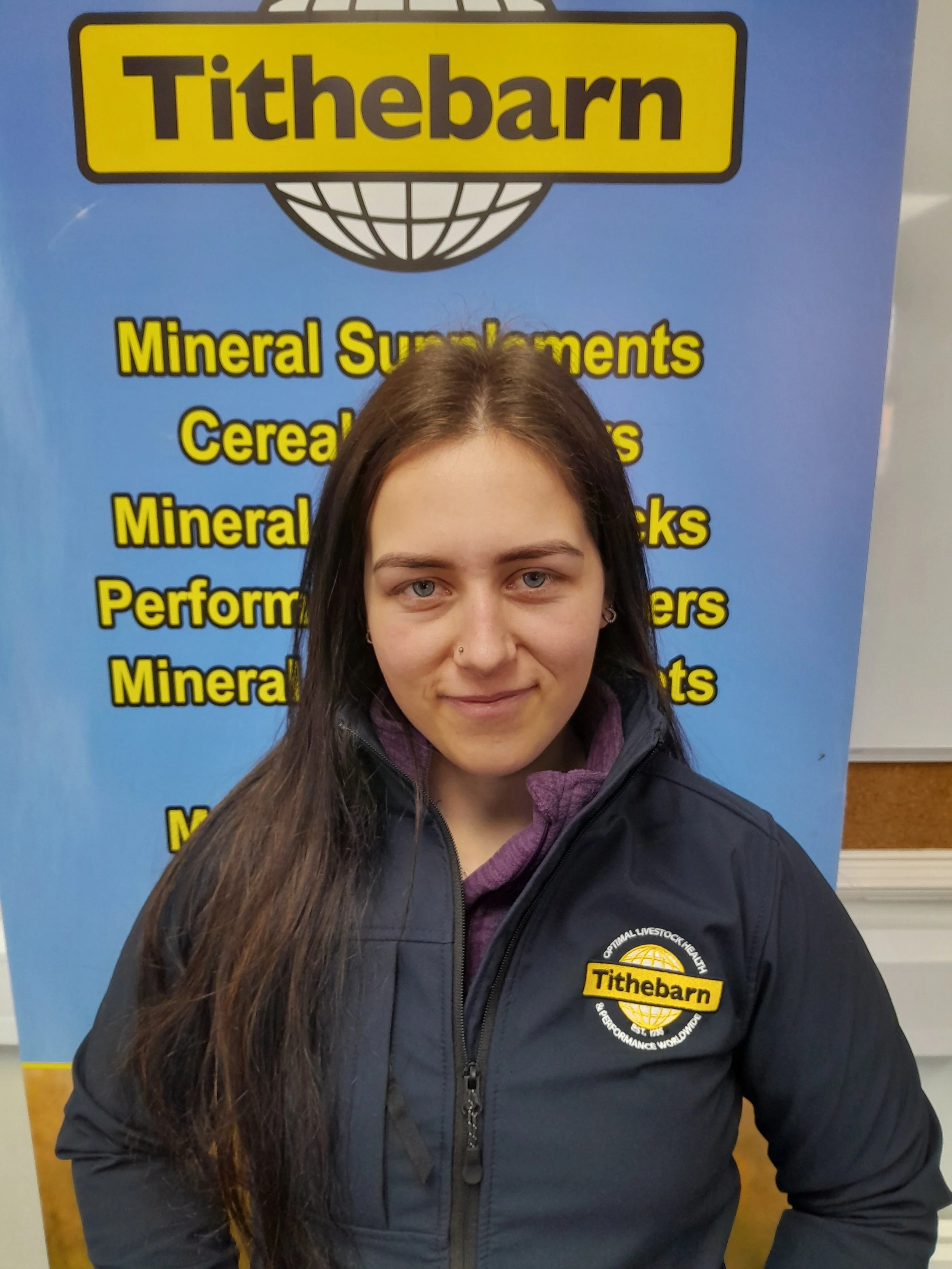 A woman with long dark hair, light skin, and gray eyes, wearing a black jacket with a yellow 'Tthebarn' logo, standing in front of a blue sign with yellow text about minerals, supplements, and performance nutrition.