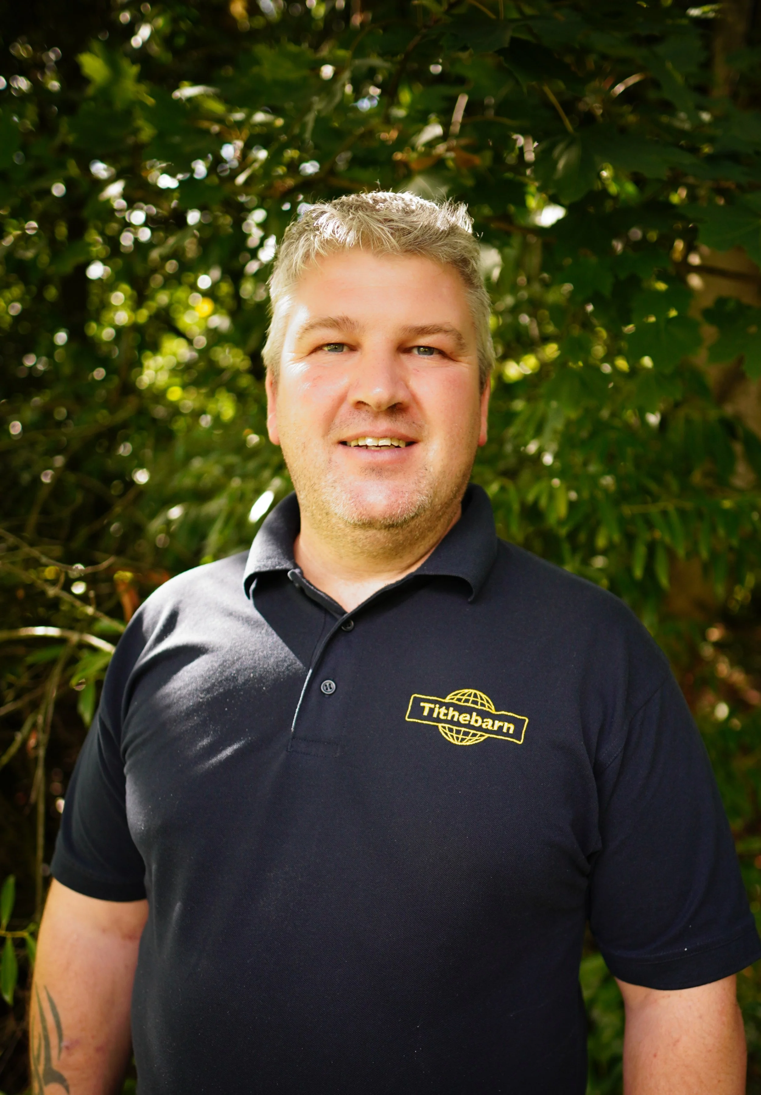 Man with short blonde hair wearing a black polo shirt with a yellow logo reading 'Tithebarn' stands outdoors amidst green foliage, smiling at the camera.