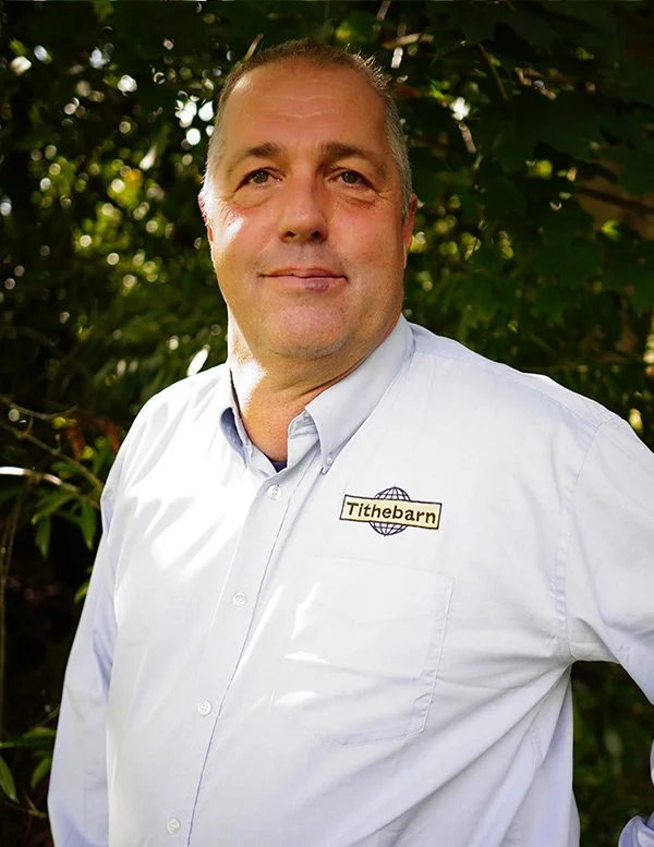A middle-aged man with short gray hair, wearing a white collared shirt with a name badge that reads "Tithebarn" and features a globe logo, standing outdoors in front of green foliage.