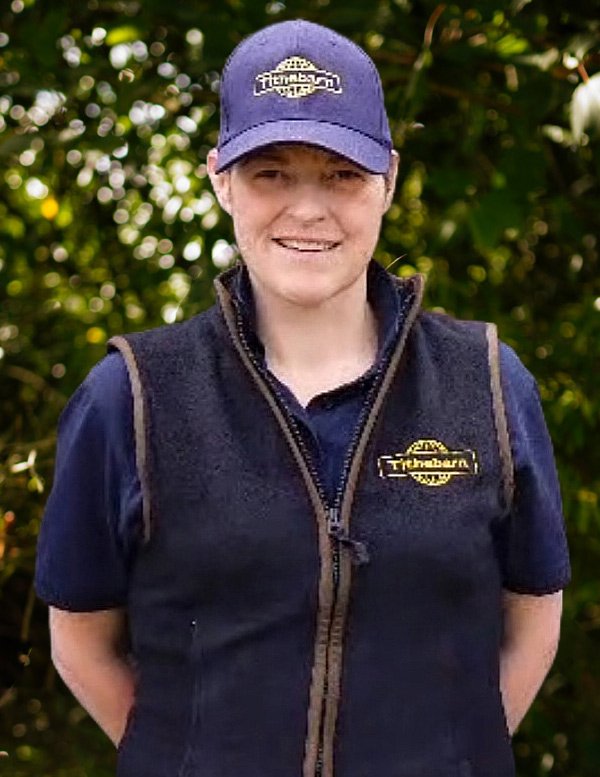 A young woman smiling outdoors, wearing a blue cap and a black vest with a logo, in front of green foliage.