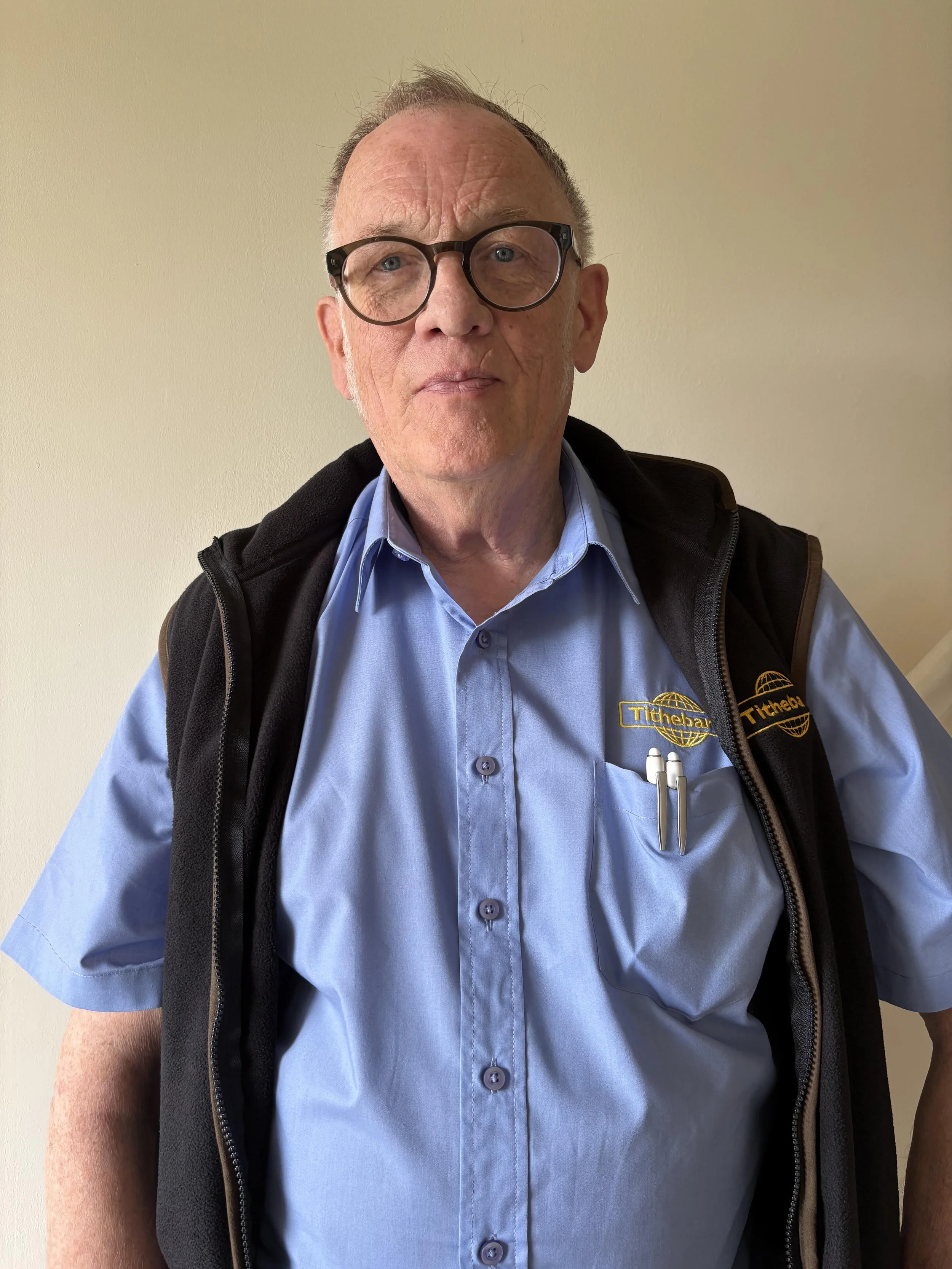 A man wearing black glasses, a light blue collared shirt, and a black vest with a logo, standing against a plain beige wall.