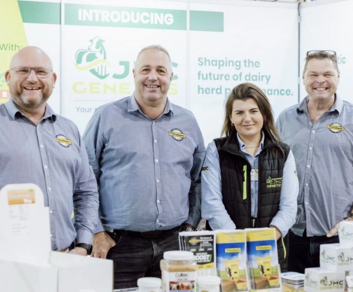 Four people standing behind a table at a trade show, three men and one woman, all wearing branded shirts and smiling. Behind them is a banner with text and logos related to dairy herd management.