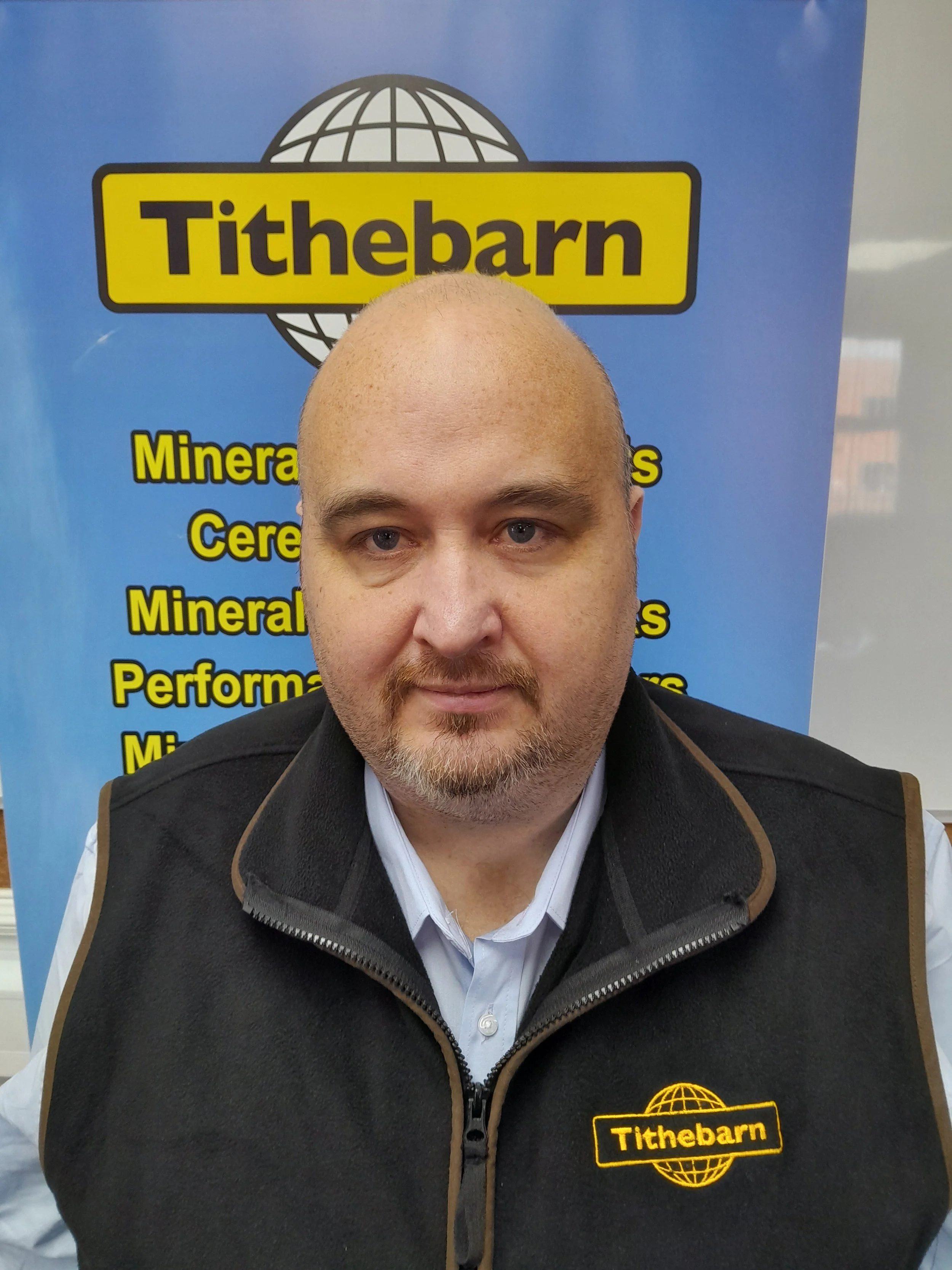 A man with a bald head and a beard wearing a black vest with a yellow Tithebarn logo on the chest, standing in front of a blue Tithebarn banner with yellow and black text.