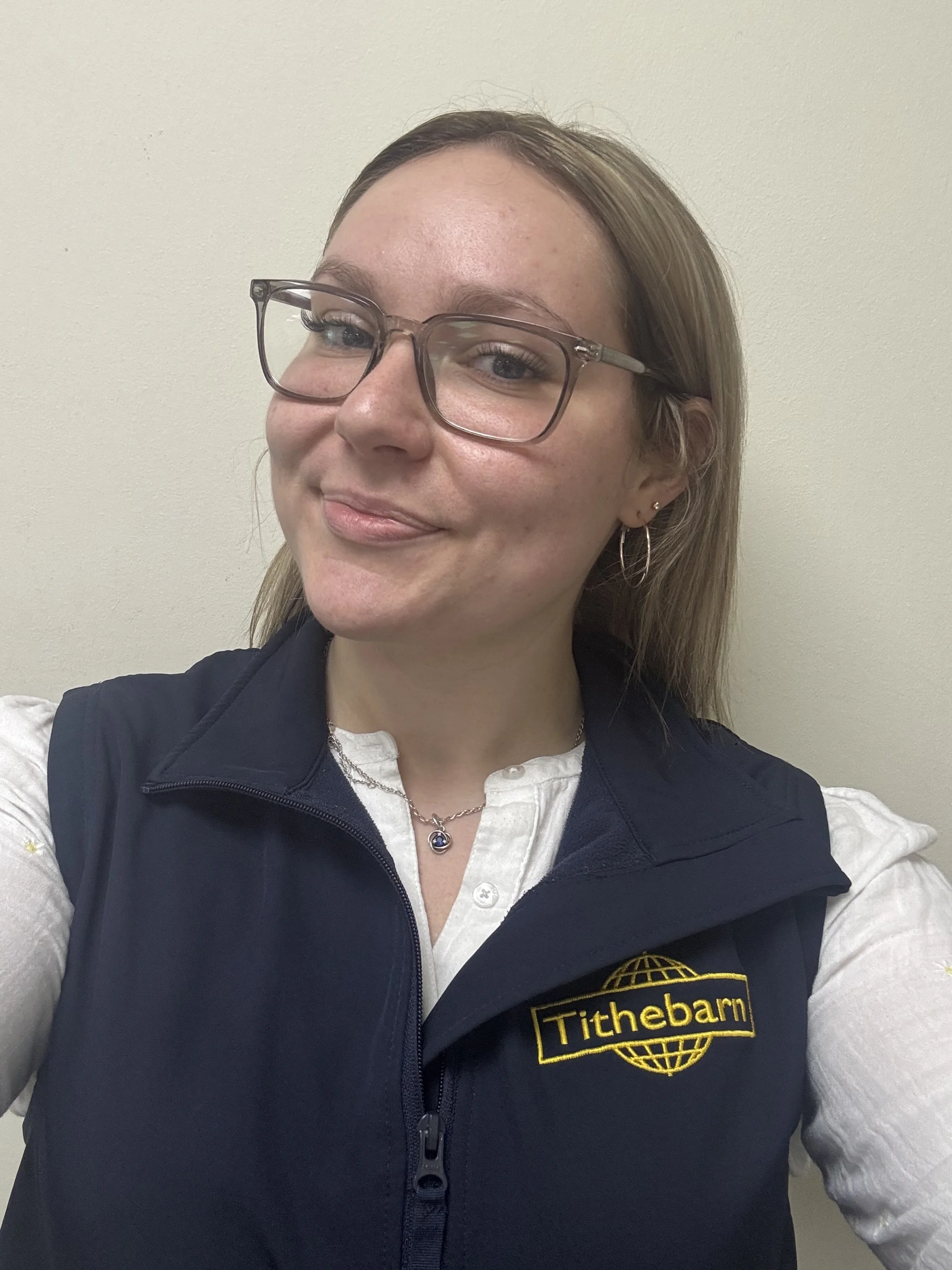A woman wearing glasses, a white shirt, and a black vest with a 'Tithebarn' logo, smiling and standing against a plain light-colored wall.