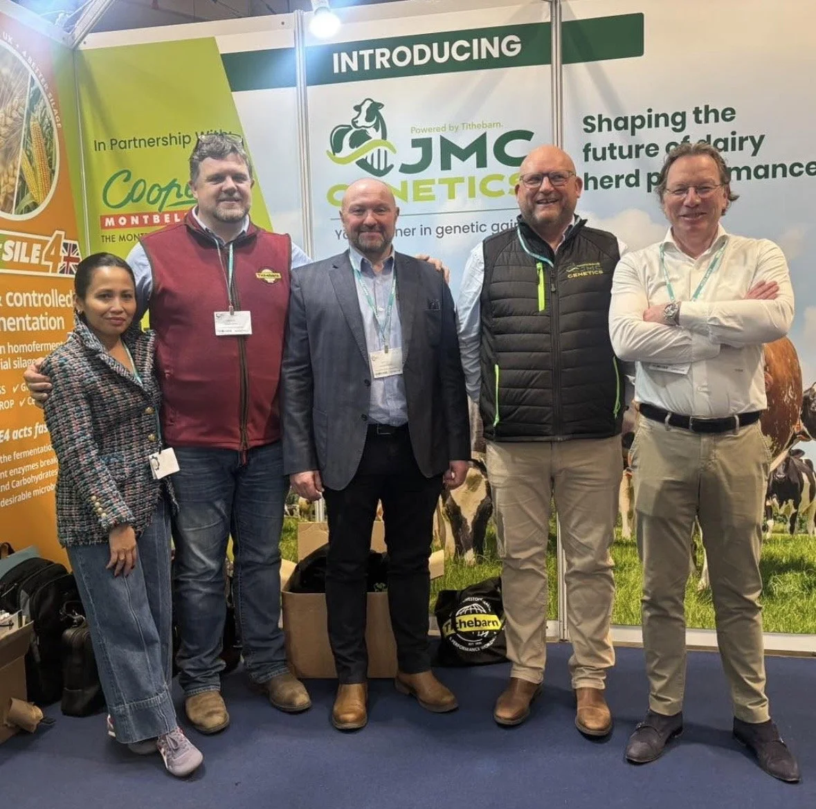 Five people standing in front of JMC Genetics booth at an event, smiling. The booth has banners about dairy herd performance and genetic gain, with images of cows and farm fields.