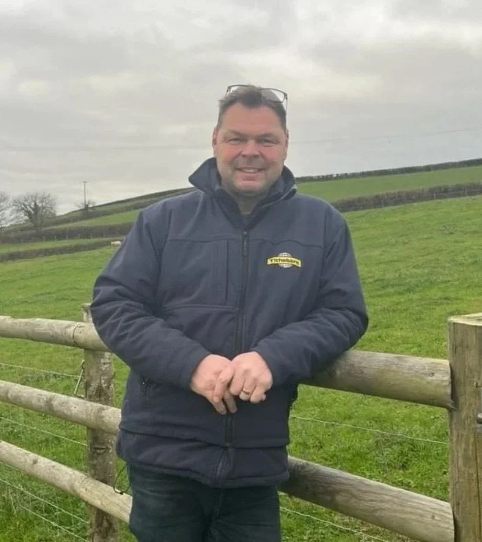 A smiling man wearing glasses on his head and a dark blue jacket standing by a wooden fence in a green field with cloudy sky.