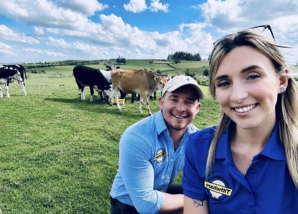 Two smiling employees in blue shirts taking a selfie in a green field with cows grazing in the background.