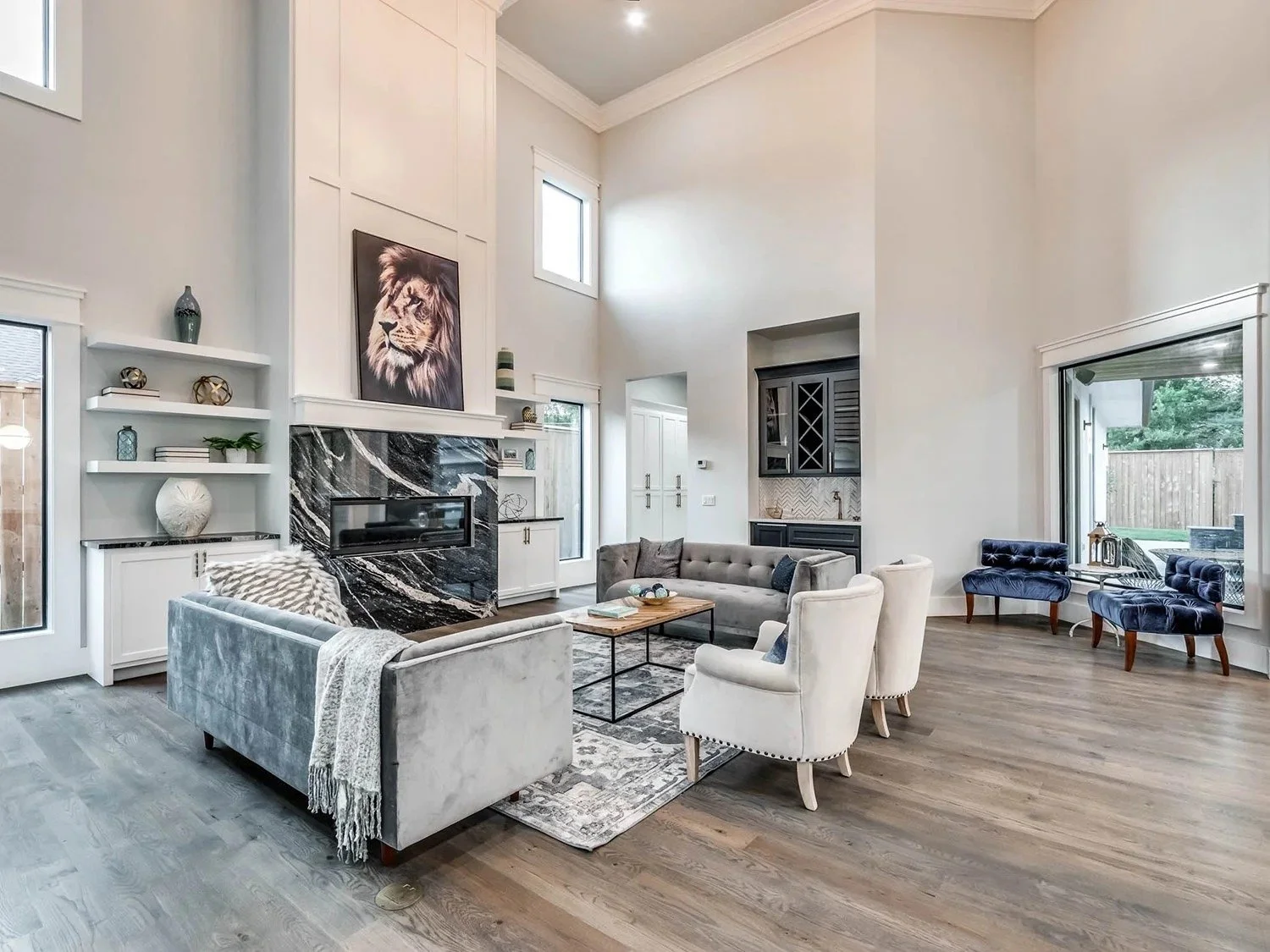 Spacious living room with high ceilings, large windows, and modern furniture including a gray sofa, white armchairs, and a black fireplace with a marble surround. Decor includes artwork of a lion and various decorative objects.