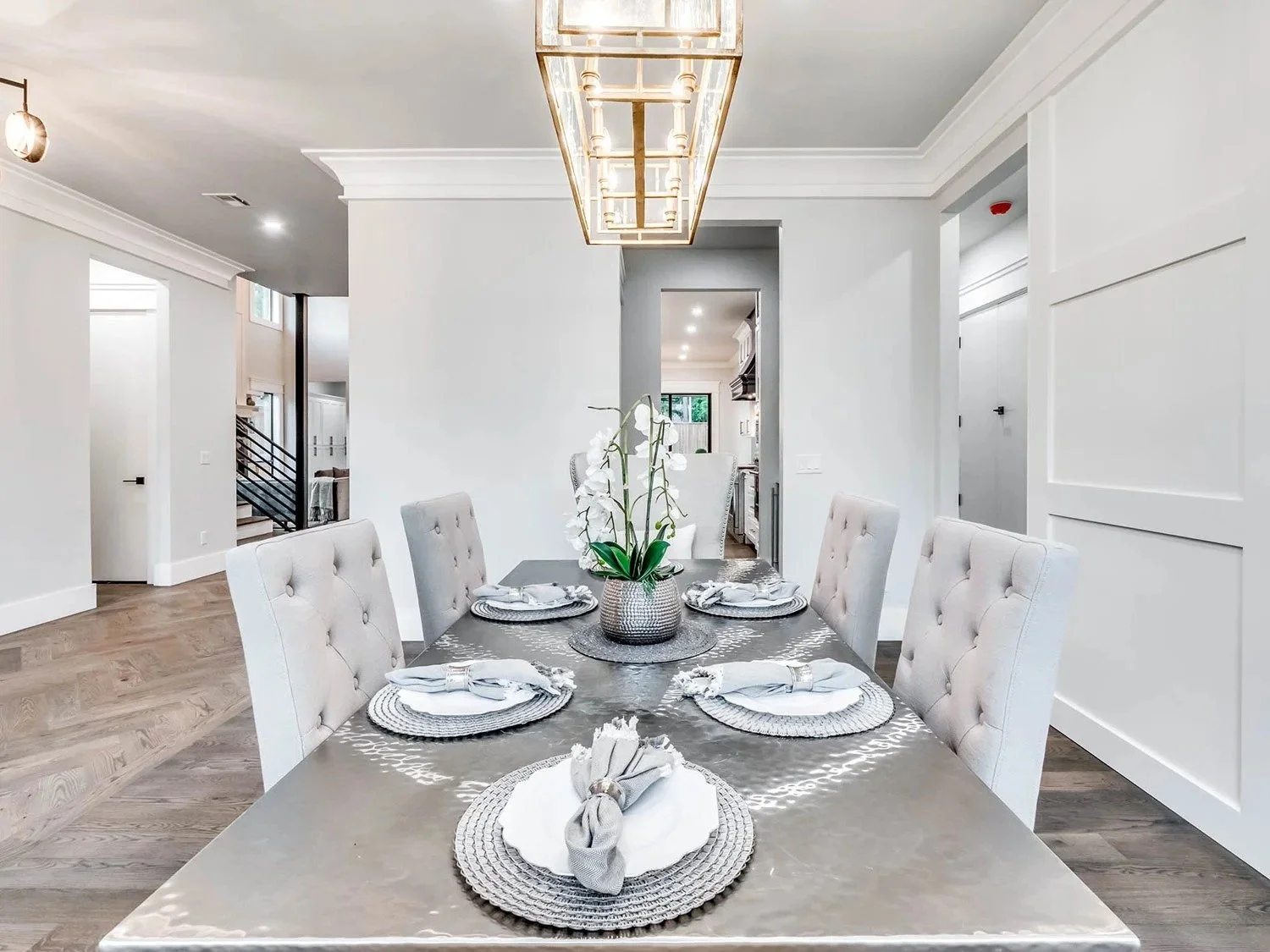 Dining room with a rectangular table set for six, with gray chairs and a vase of white orchids as a centerpiece, in a modern home with white walls and hardwood flooring.