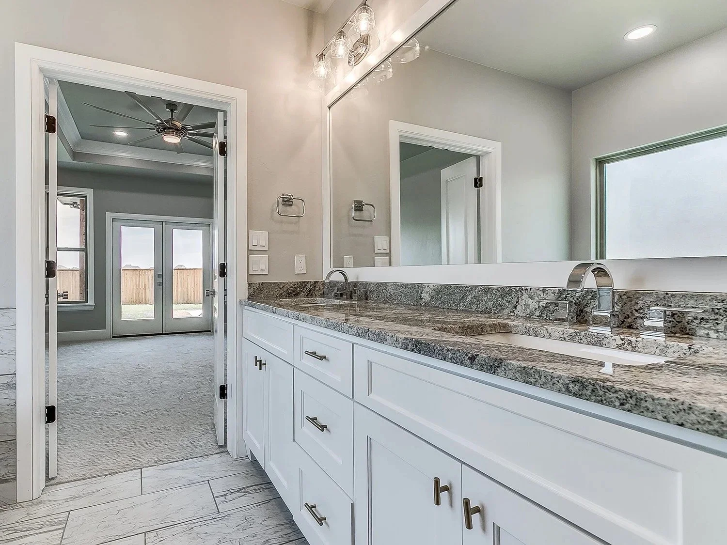 Modern bathroom with white cabinets, granite countertops, large mirror, and dual sinks. View into an adjacent room with ceiling fan and outside doors.