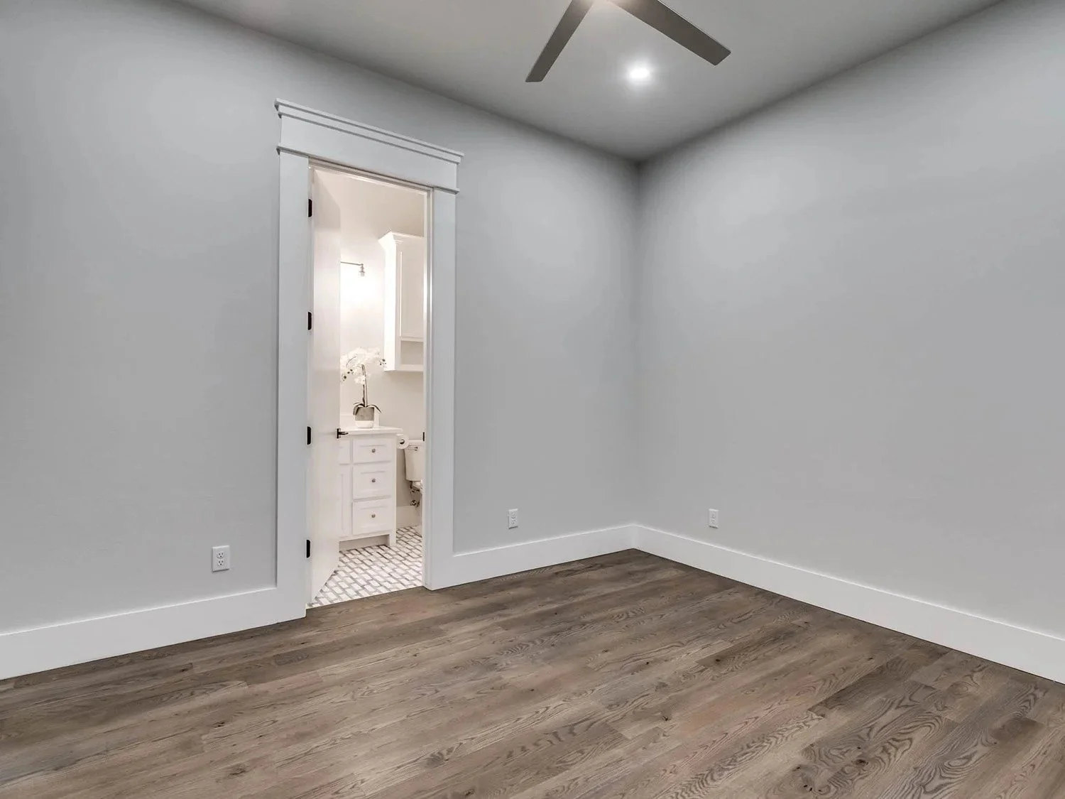 Empty room with hardwood flooring, white painted walls, and an attached bathroom visible through an open door.