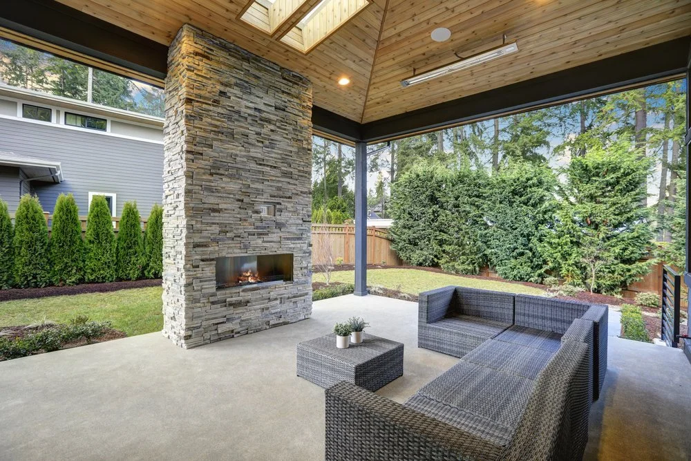 Covered outdoor patio with wicker sectional sofa, a small coffee table with potted plants, stone fireplace, and large windows showing a green backyard with trees and bushes.