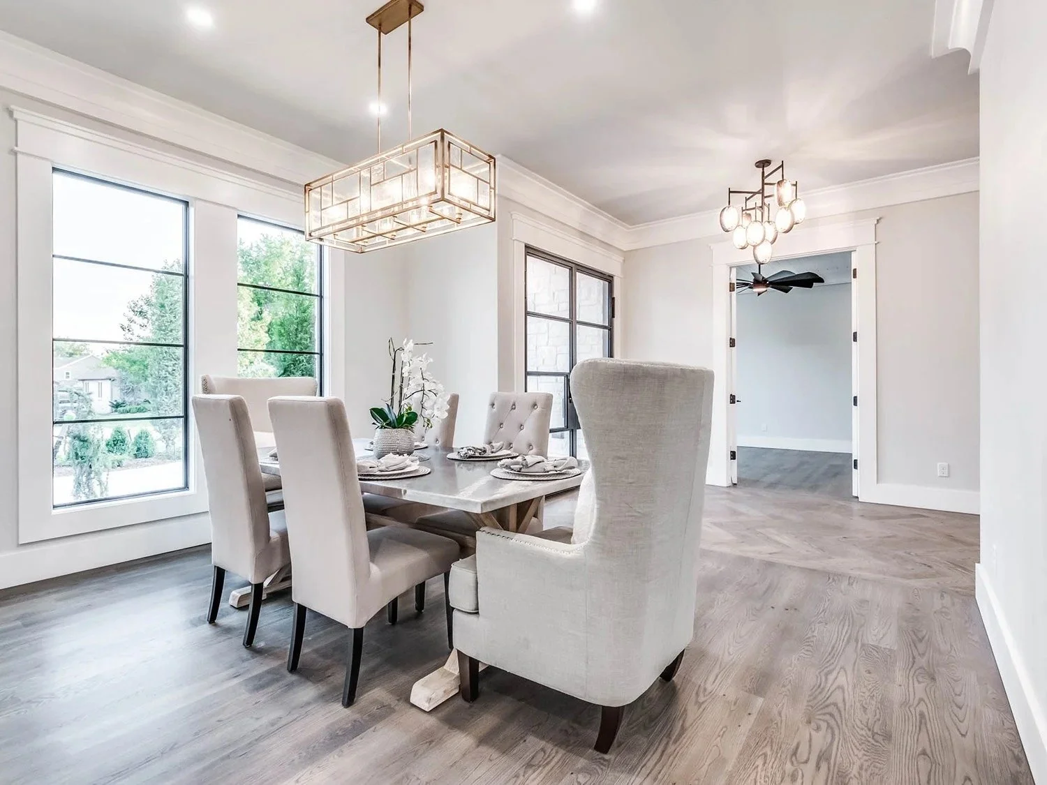Bright dining room with large windows, white walls, light wood flooring, a rectangular dining table, and upholstered chairs, decorated with a flower arrangement, pendant lighting, and a chandelier.