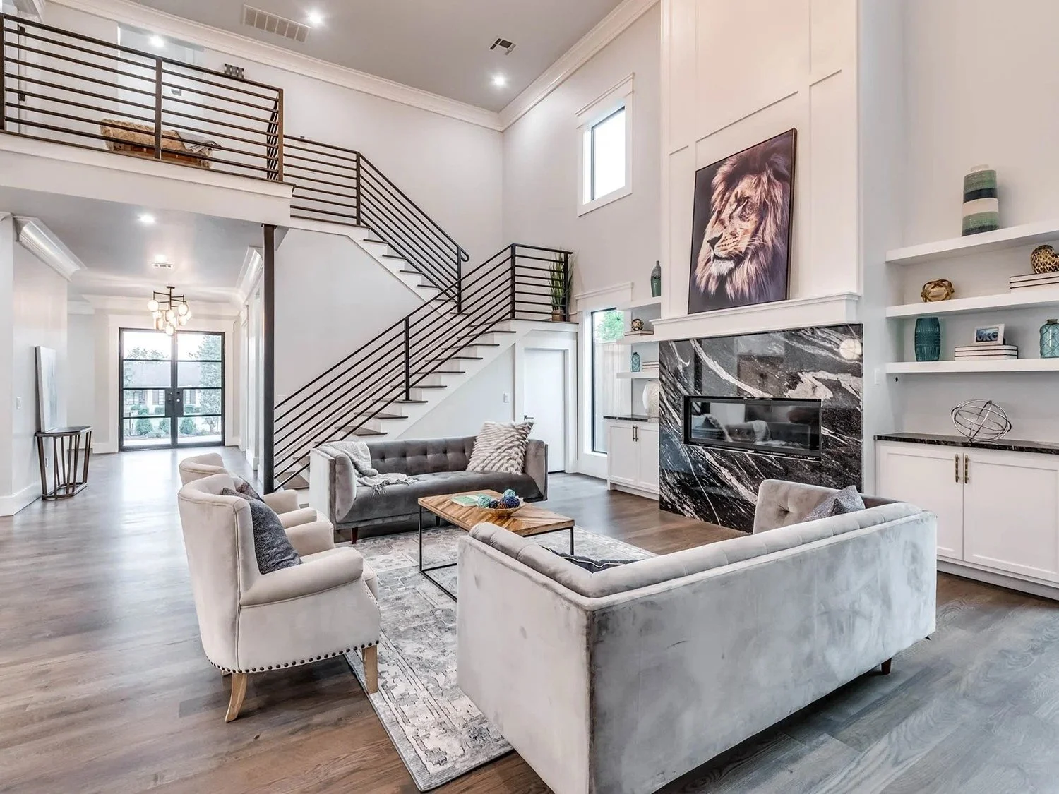 Spacious living room with modern decor, including a gray velvet sofa, white armchairs, a wooden coffee table, and a fireplace with black marble surround. Tall ceilings and a staircase with black metal railings lead to an upper level. Built-in white s