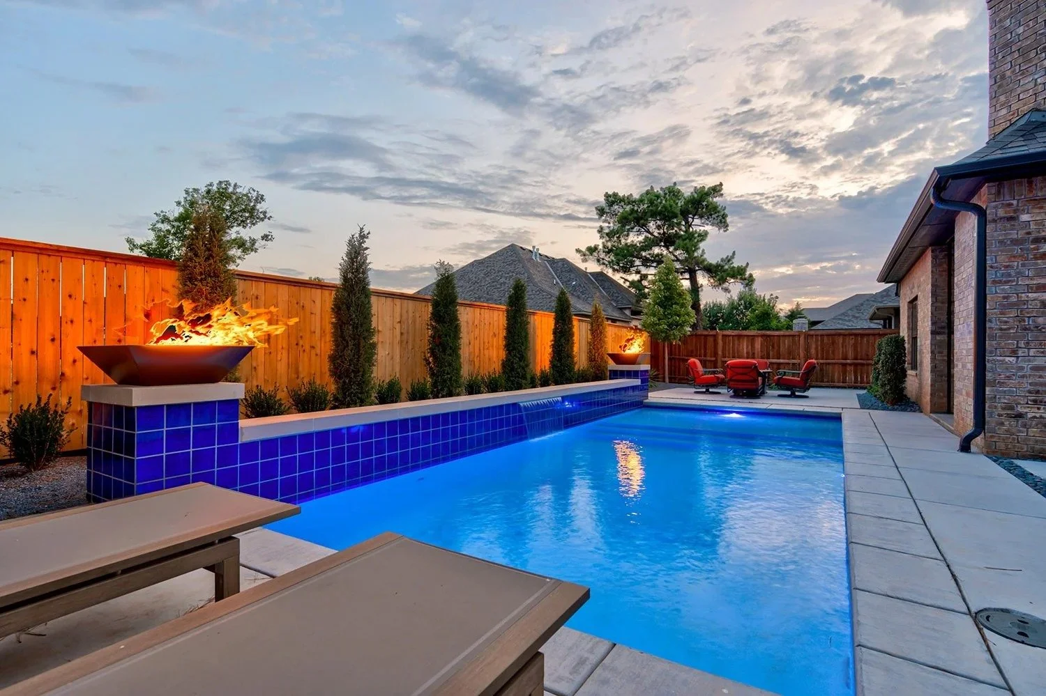 Backyard with a swimming pool, wooden fence, tall trees, and outdoor furniture at sunset.