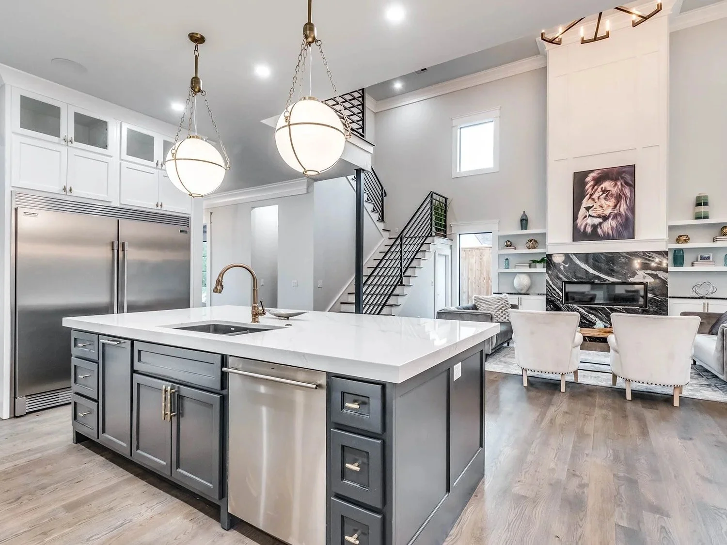 Modern kitchen with a large island, stainless steel appliances, white cabinetry, and a living room with white chairs and a fireplace with a lion portrait above it.