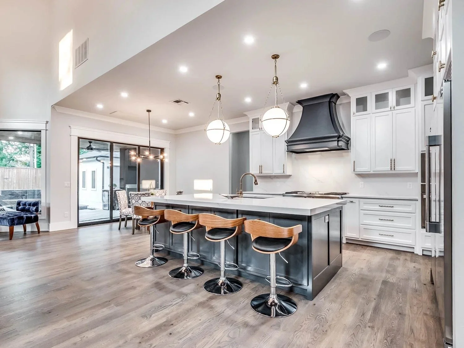 Modern kitchen with white cabinets, a large island with a dark base, four wooden bar stools, and two hanging pendant lights. There is a dining area with a table and chairs, sliding glass doors, and hardwood flooring.