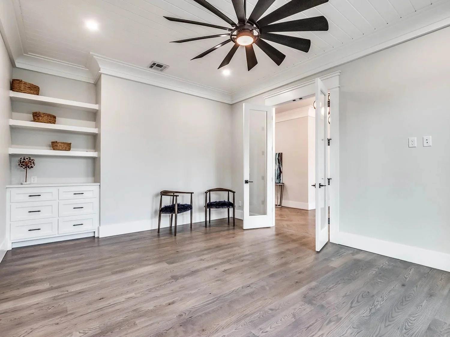 An empty room with light-colored walls and hardwood flooring, two chairs near a doorway, built-in shelves with baskets, a ceiling fan, and a doorway leading to another room