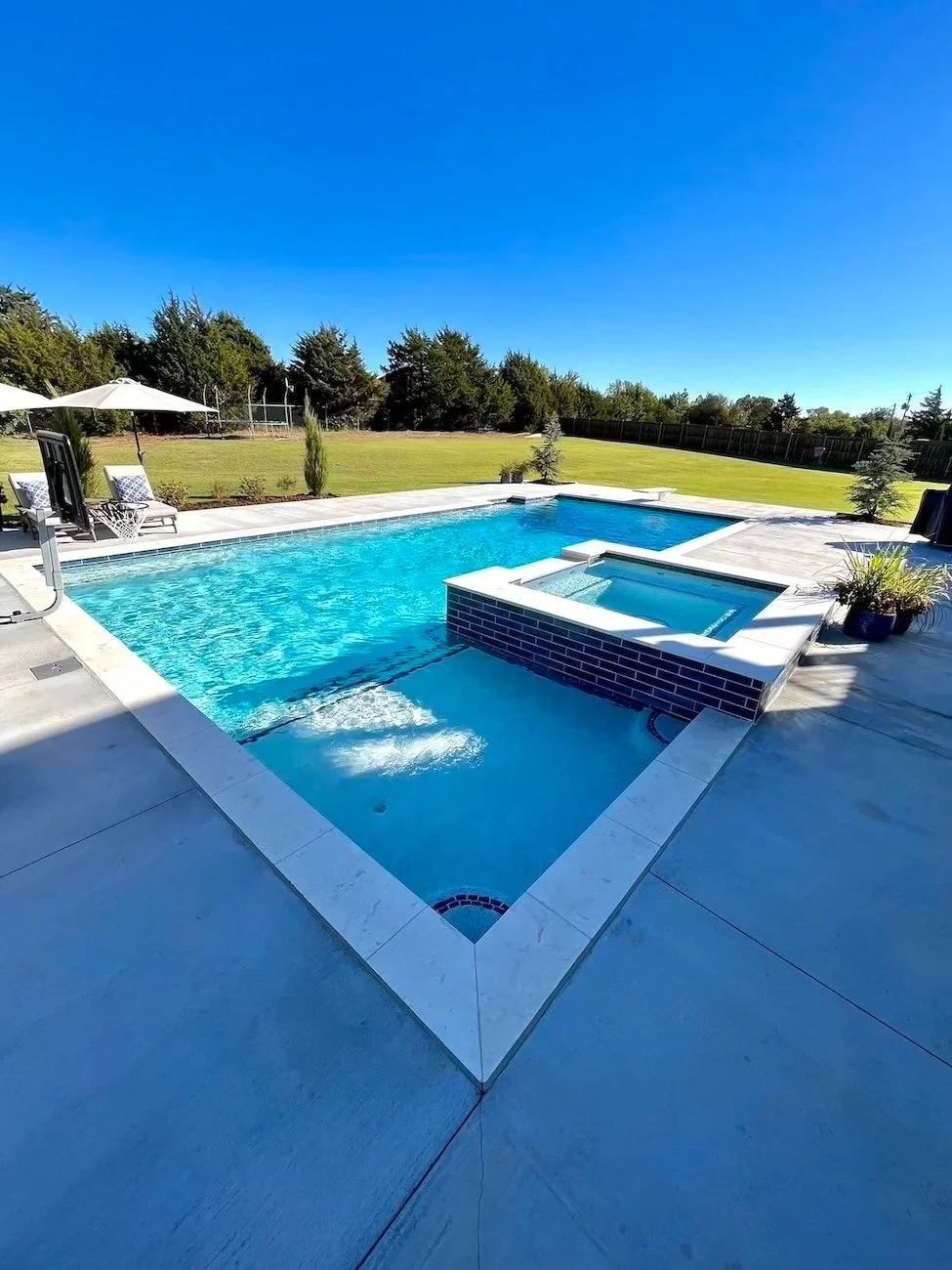 Backyard with a rectangular swimming pool and a hot tub, surrounded by a white concrete deck, with chairs and umbrellas on the side, and a large grassy area with trees under a clear blue sky.