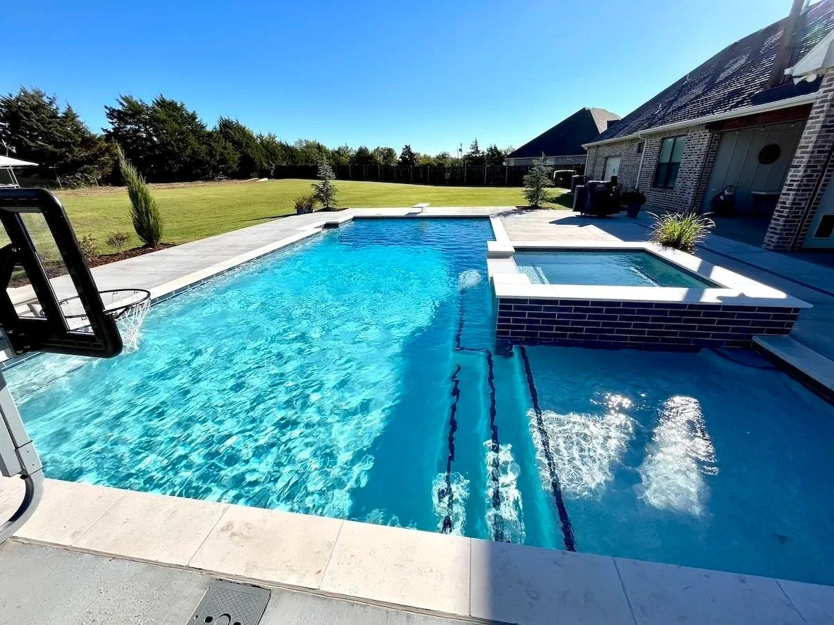 Backyard with a rectangular swimming pool, a hot tub with brick exterior, and a grassy yard with trees in the background under a clear blue sky.