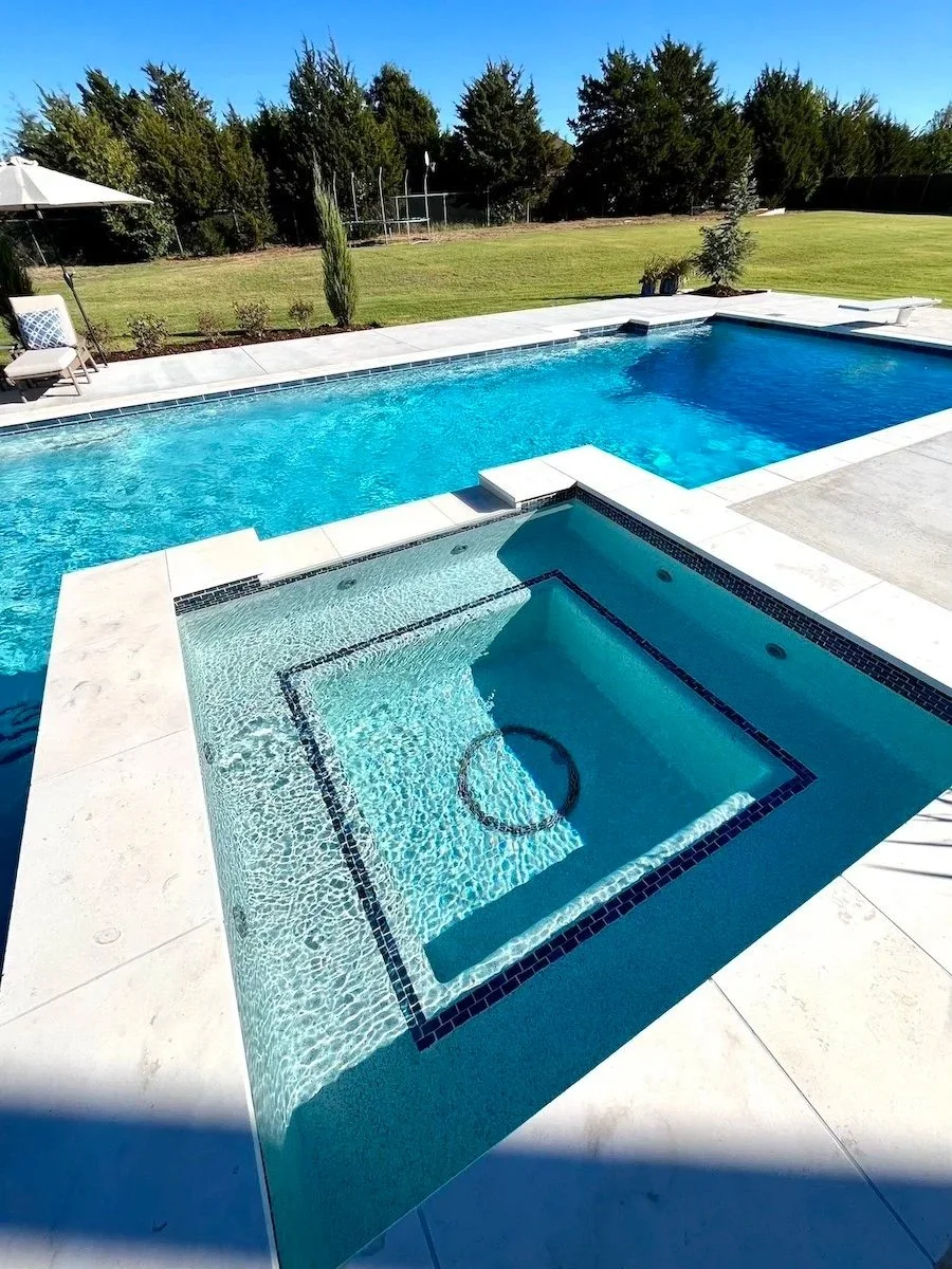 A backyard with a large in-ground swimming pool and a hot tub, surrounded by a white stone deck, with lounge chairs, a large umbrella, and a grassy area with trees and shrubs in the background.